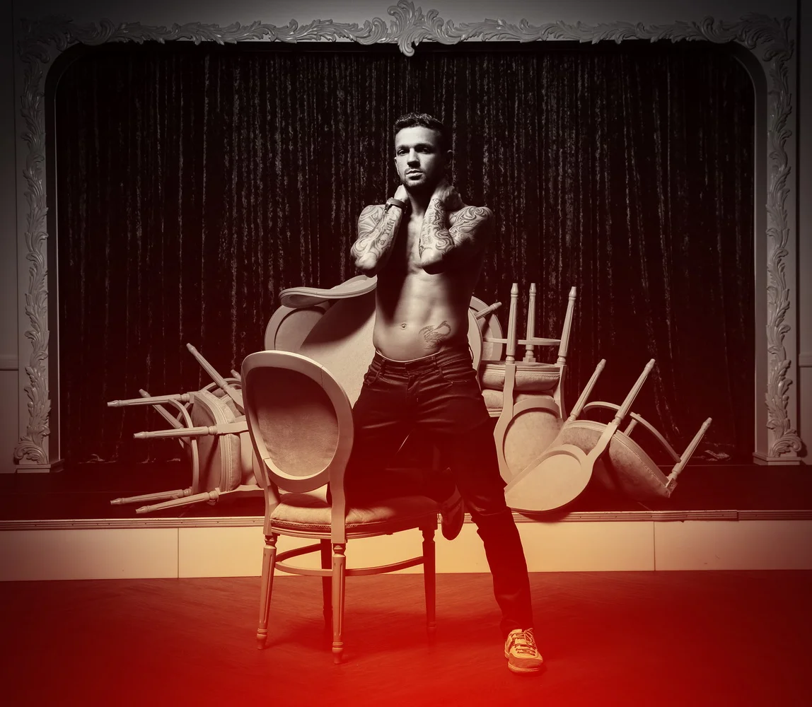 A shirtless man with tattoos standing on a stage with overturned chairs and a dark curtain backdrop.