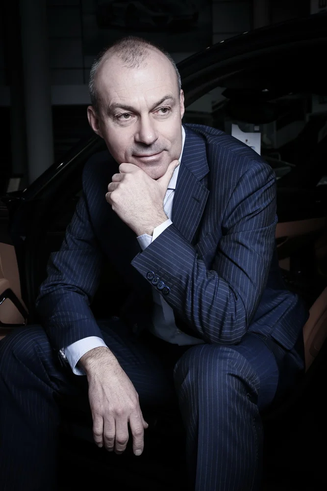 A middle-aged man in a dark pinstripe suit sitting on a car seat, resting his chin on his hand, looking thoughtful.