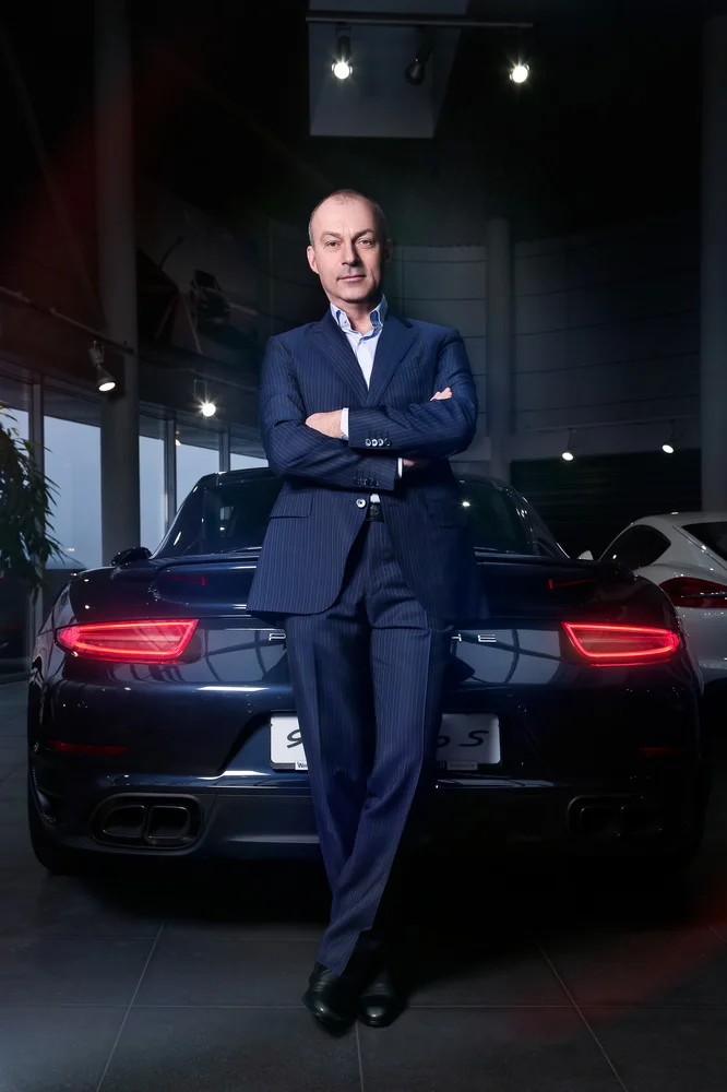 A man in a navy pinstripe suit standing with arms crossed in front of a black luxury car inside a showroom.