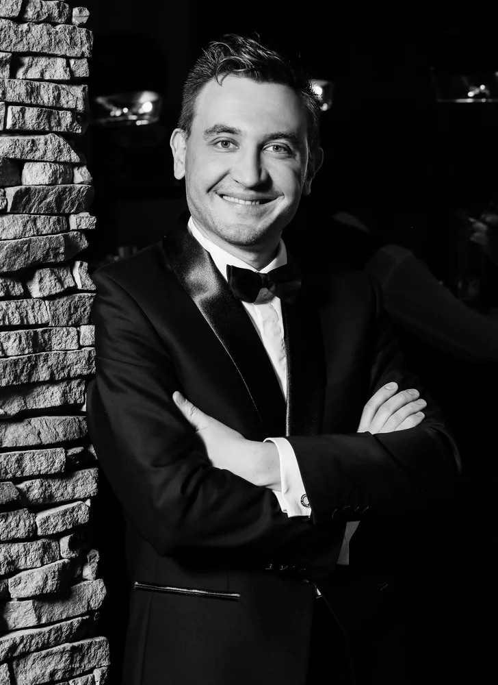 Black and white photo of a man in a tuxedo with arms crossed, smiling, standing next to a stone wall.
