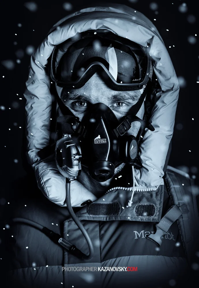 Close-up of an ice climber wearing a helmet, goggles, and oxygen mask, with snow falling around him.