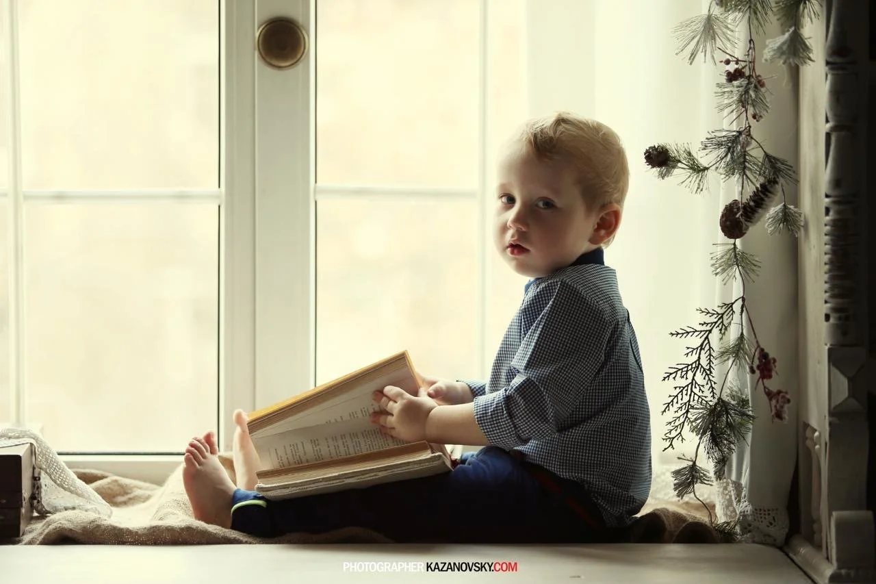 A young child sitting on a window seat, holding an open book, with a window and decorative plant in the background.