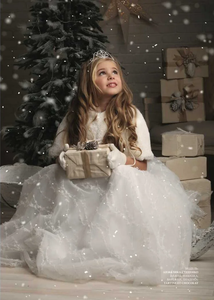 A young girl dressed as a princess in a white gown with a tiara, sitting on the floor holding a wrapped gift box with a pinecone ornament. She is in a festive Christmas setting with a decorated Christmas tree and wrapped presents in the background, a