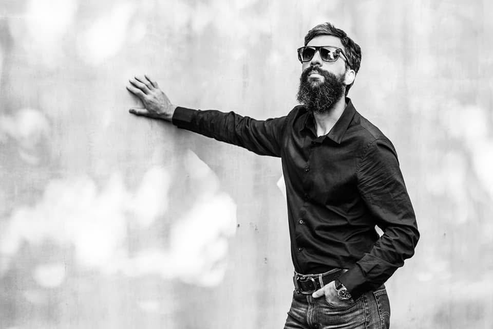 A man with a beard and sunglasses wearing a black shirt and jeans standing against a plain wall, with his hand resting on the wall.
