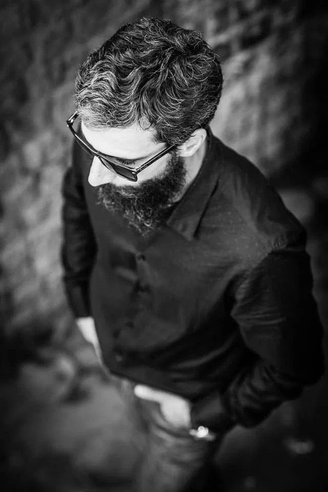 Black and white photo of a man with glasses, curly hair, and a beard, looking down. He is wearing a dark jacket and has one hand in his pocket.