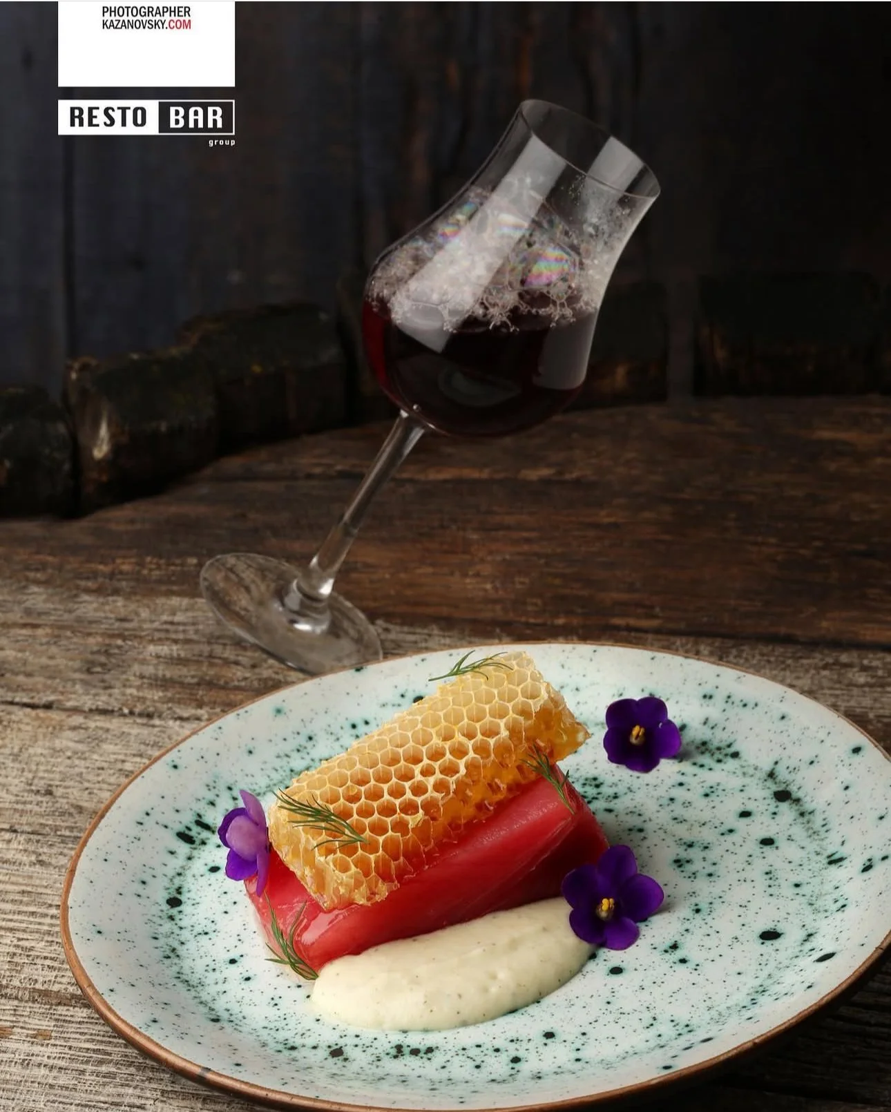 A culinary dish featuring a piece of cured meat, topped with honeycomb, garnished with purple edible flowers and dill, served with a creamy white sauce on a speckled ceramic plate, with a glass of red wine in the background.