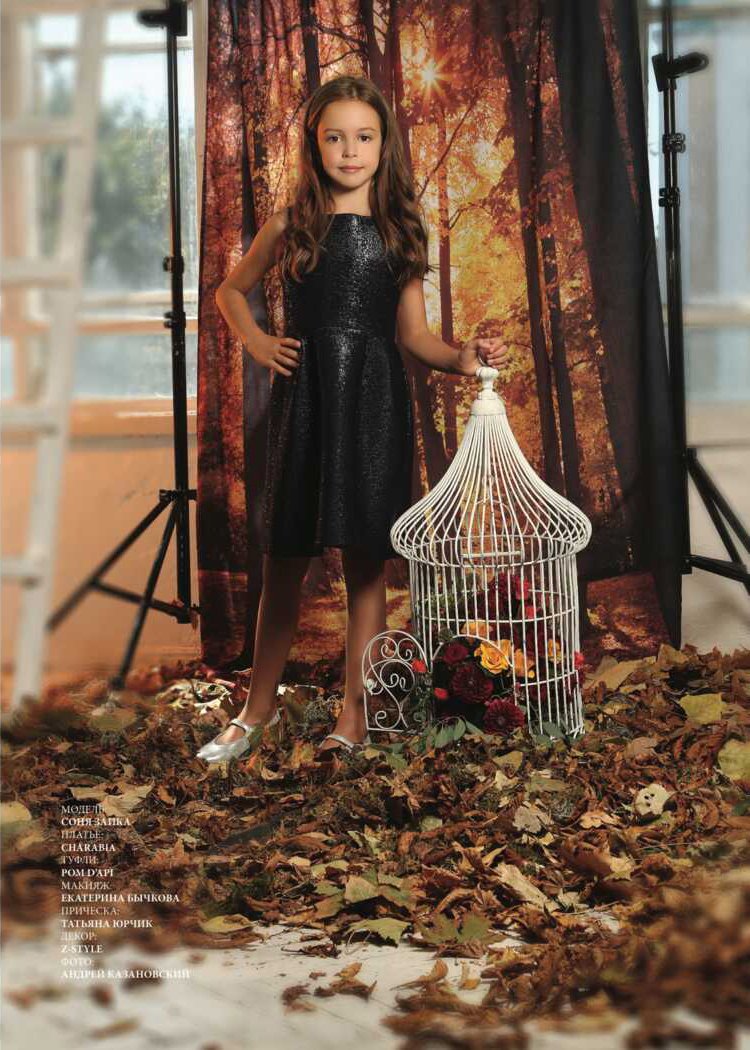 A young girl with long brown hair wearing a black, sparkly dress and silver shoes standing in front of a woodland scene backdrop, holding a white birdcage filled with colorful roses, with autumn leaves scattered on the ground.
