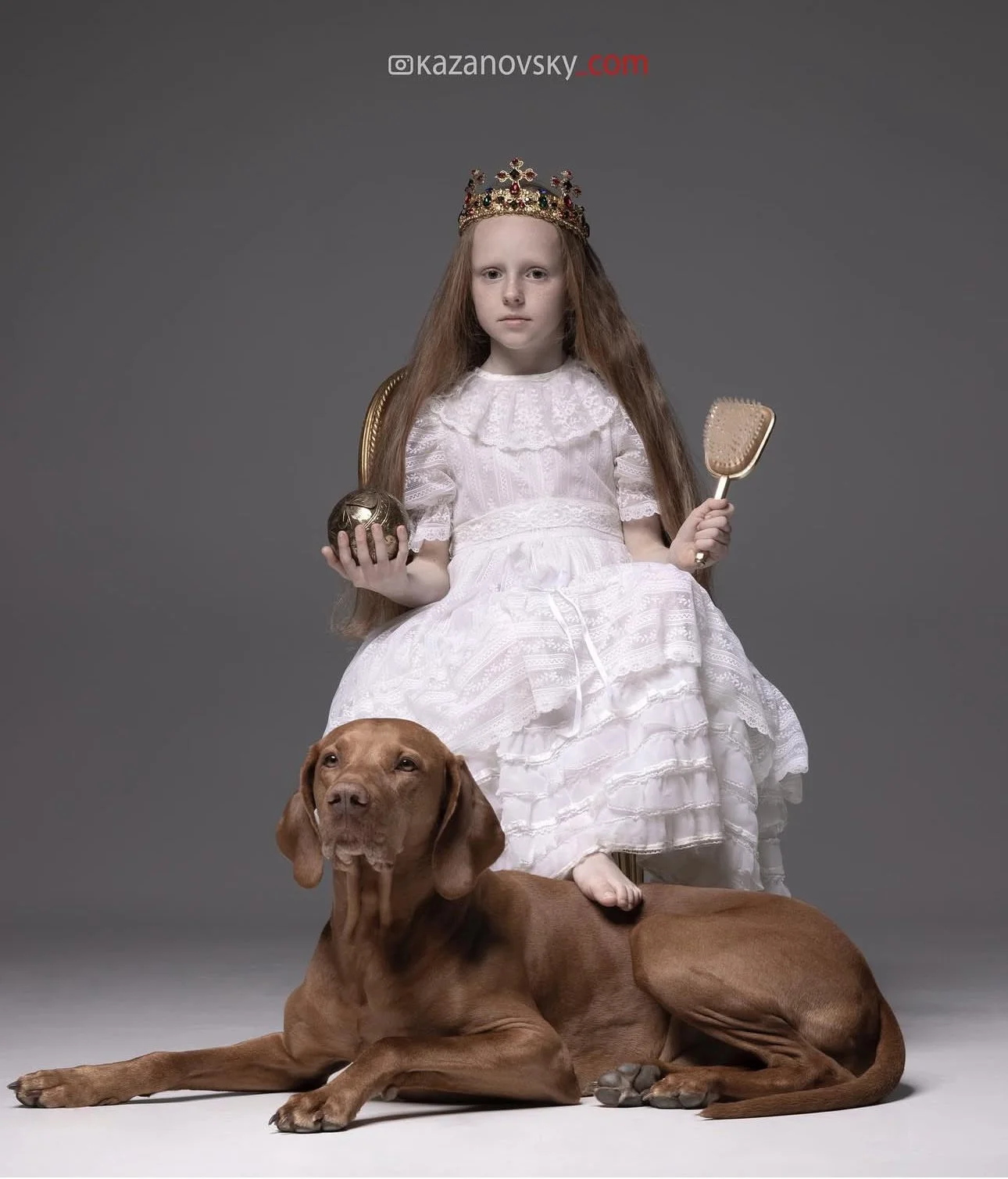 Young girl dressed as a queen with a crown, sitting on a chair with a golden backrest, holding a globe in one hand and a brush in the other. A large brown dog is lying on the floor in front of her, with one paw on her lap, against a plain gray backgr