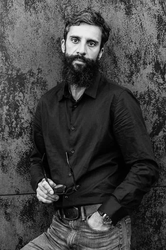 A man with a beard and short hair, wearing a black button-up shirt and jeans, standing against a textured wall, holding eyeglasses in one hand and wearing a wristwatch.