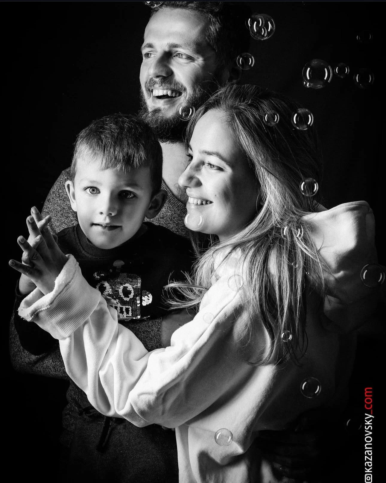 A black and white photo of a happy family with two adults and a young boy, smiling and embracing each other, surrounded by floating bubbles.
