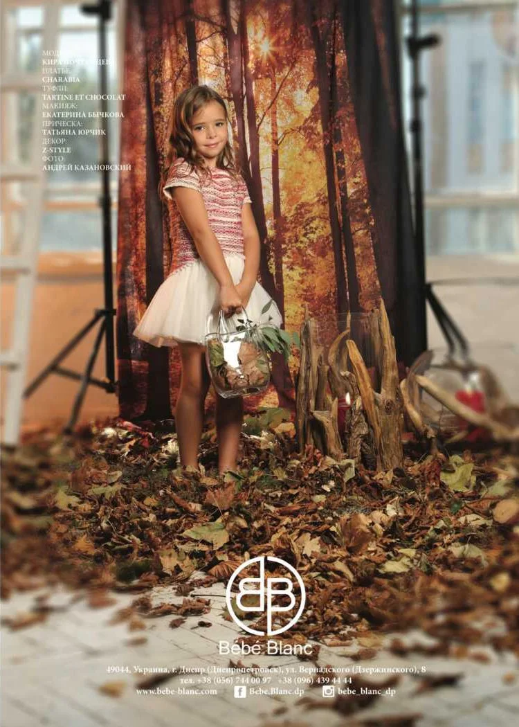 A young girl standing on a leaf-covered floor, holding a glass basket with leaves and branches, in front of an autumn-themed backdrop depicting a forest with sunlight filtering through trees. She is dressed in a pink patterned top and a white skirt, 