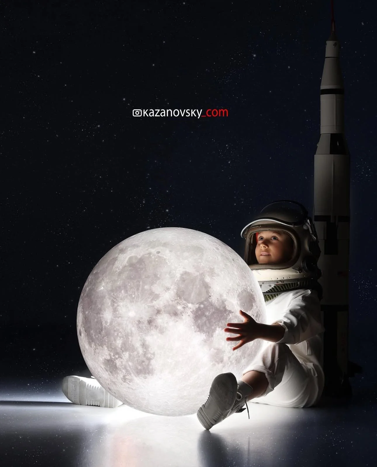 A child dressed as an astronaut sitting next to the moon with a rocket in the background, holding or touching a large illuminated globe of the moon, against a starry night sky.