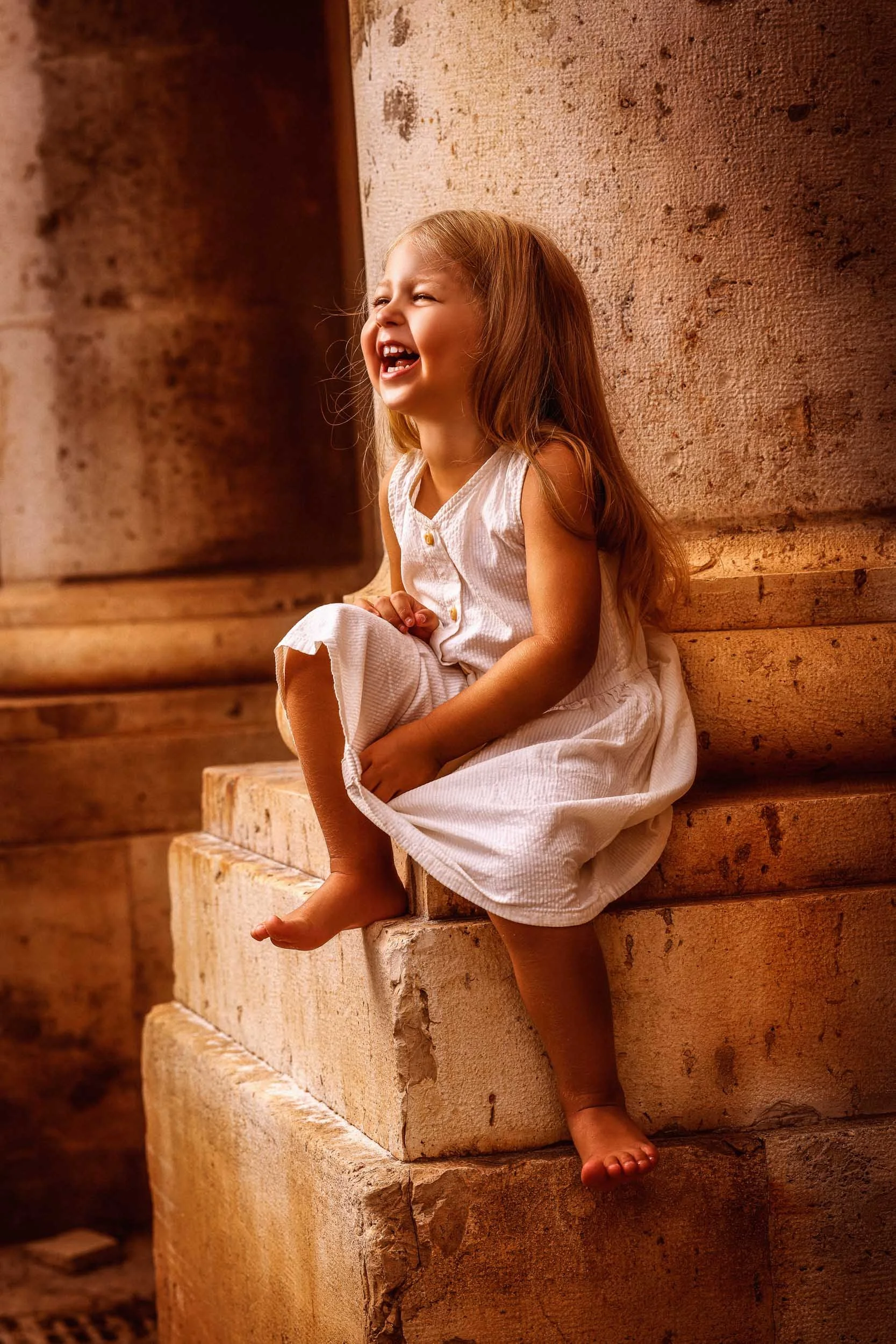 A young girl with long, light brown hair laughing while sitting on stone steps next to a textured stone wall, wearing a white sleeveless dress.