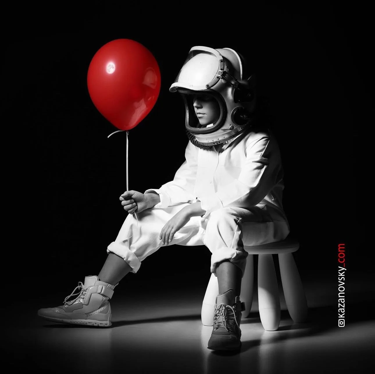 Child in space suit helmet sitting on a small stool, holding a red balloon, with a black background.