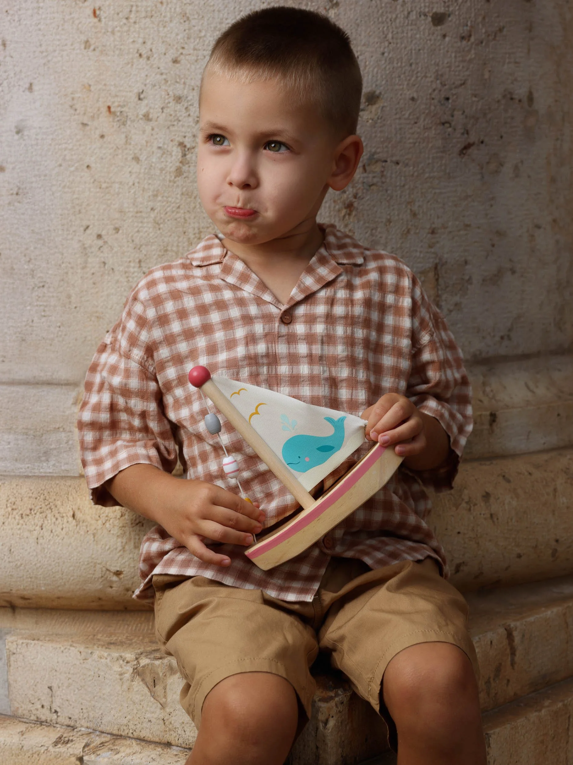 A young boy with short blond hair, wearing a checkered shirt and khaki shorts, sitting against a textured stone wall, holding a toy sailboat with a whale illustration on the sail.
