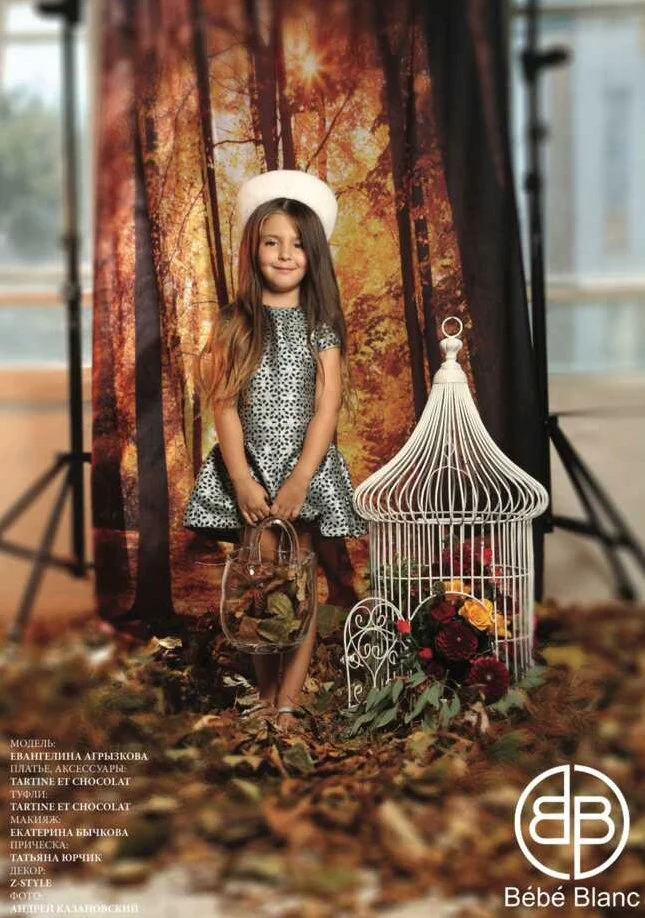 A young girl with long brown hair, wearing a black and white leopard print dress and a white hat, stands in front of an autumn forest backdrop. She is holding a small basket with leaves inside, and next to her is a white decorative birdcage with flow