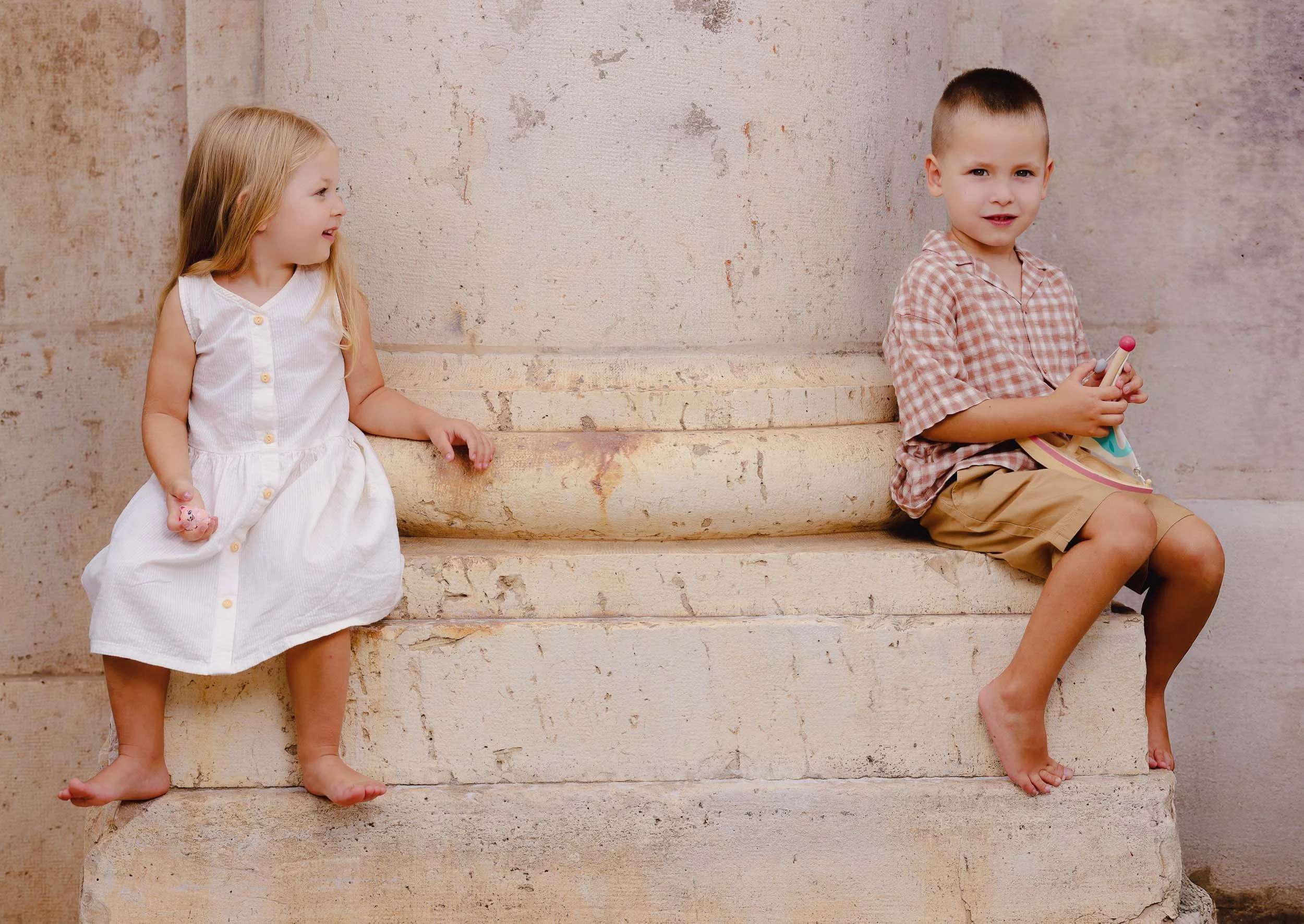 Two young children, a girl and a boy, sitting on stone steps in front of a textured beige wall. The girl has long blonde hair, is wearing a white sleeveless dress, and is smiling while looking at the boy. The boy has short brown hair, is wearing a pl