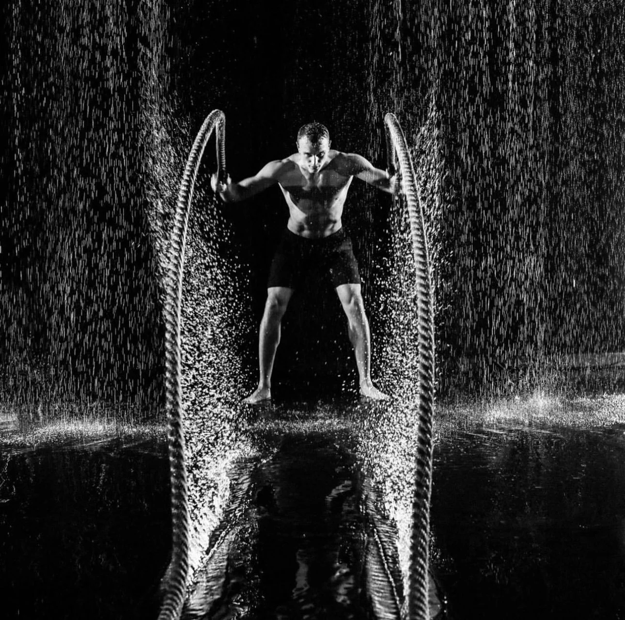 A shirtless man in shorts is standing in the center of a dark room, holding onto two ropes and water falling around him.