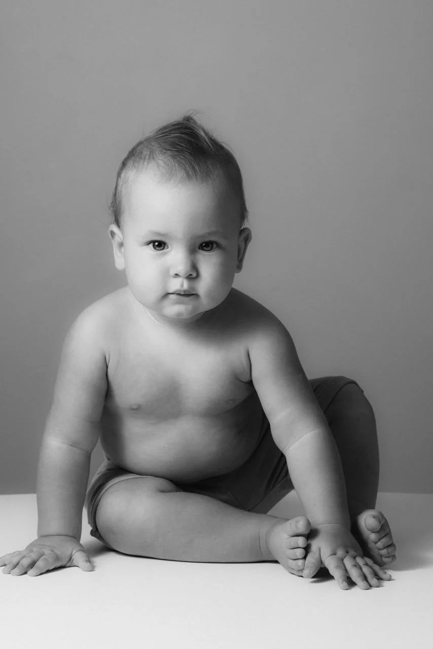 Black and white photo of a baby with short hair sitting on the floor, looking at the camera.