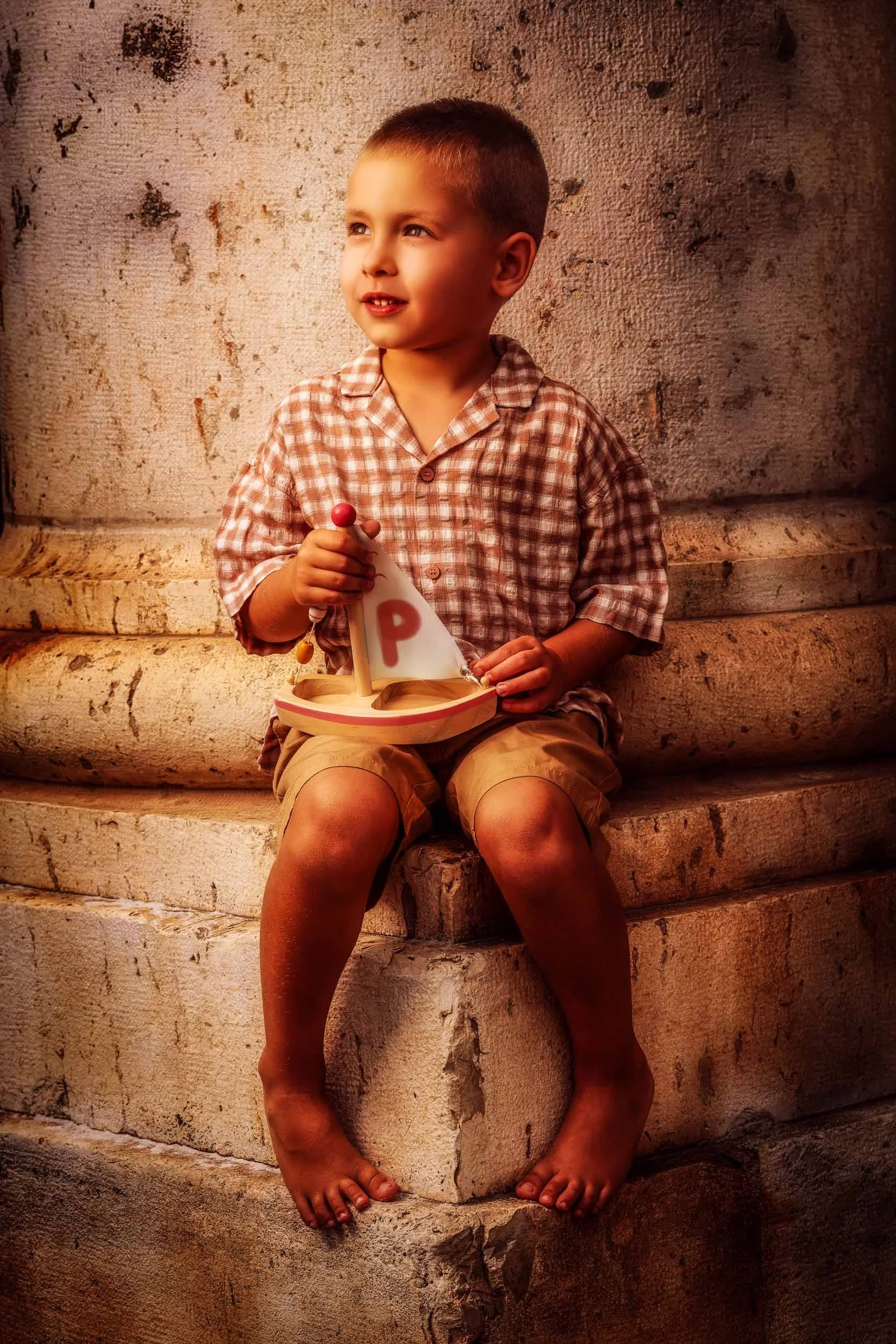 A young boy sitting on stone steps, holding a small sailboat with a white sail and a red letter P on it, looking to the side with a slight smile.