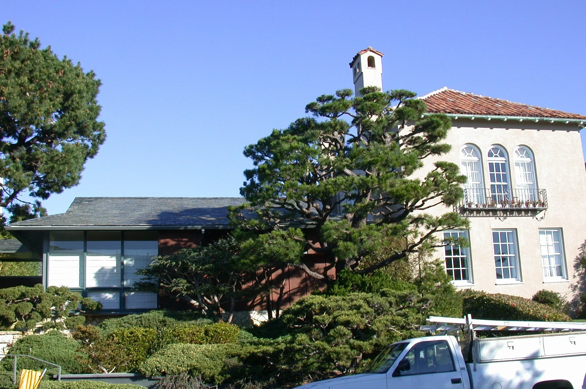 A building with a tile roof and white walls, trees in the foreground, and a pickup truck parked outside on a clear day with blue sky.