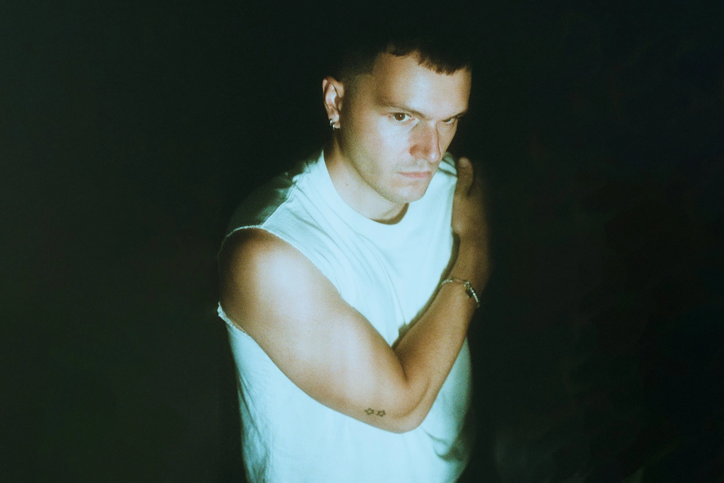 A person with short dark hair, wearing a sleeveless white shirt, looking into the camera with a serious expression, in a dark environment.