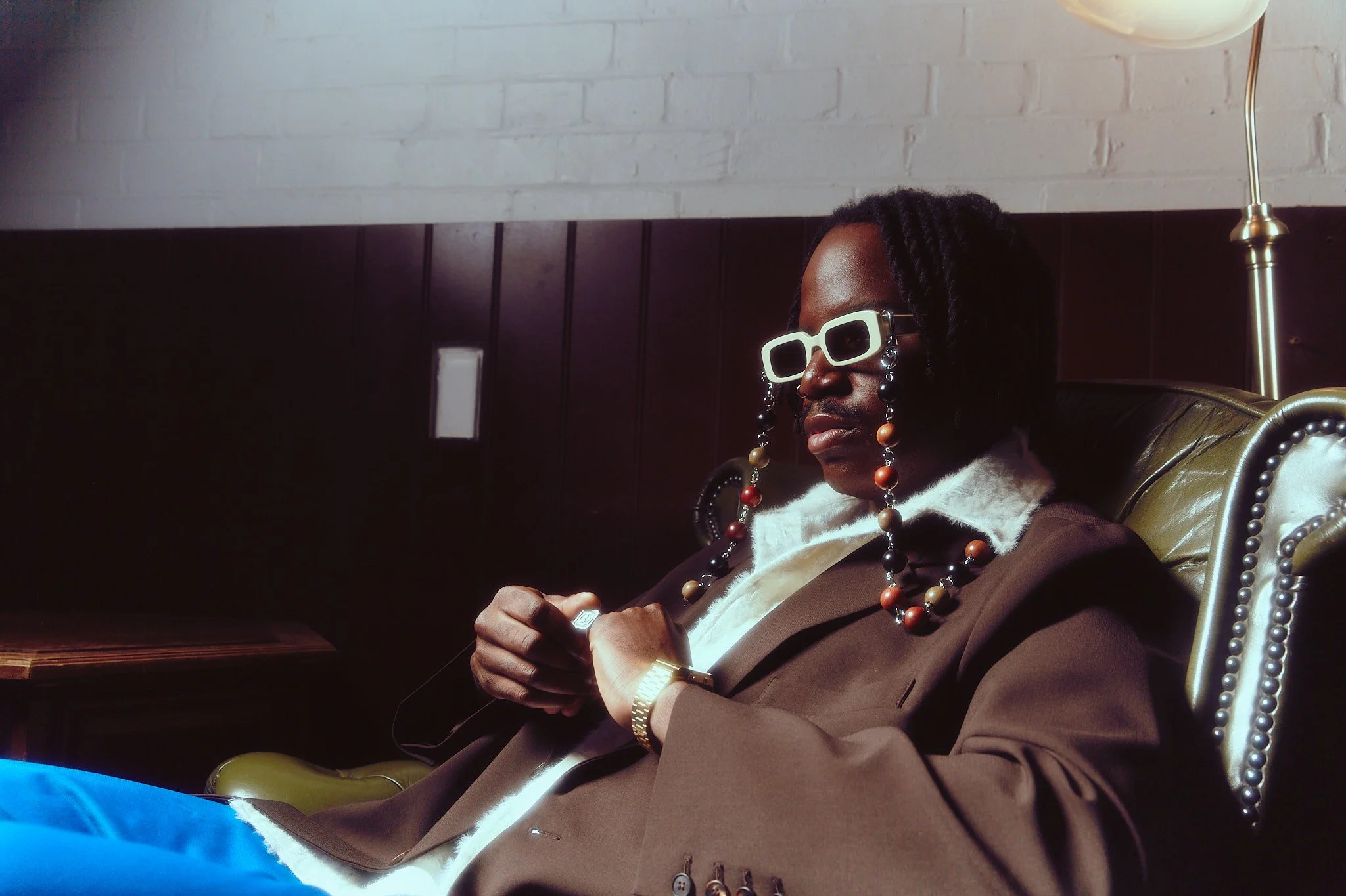 A person with dreadlocks wearing white sunglasses, colorful bead jewelry, a brown blazer, and a watch, sitting on a vintage green leather armchair against a dark wall with a white brick upper section, holding a cigarette in a room with subdued lighti