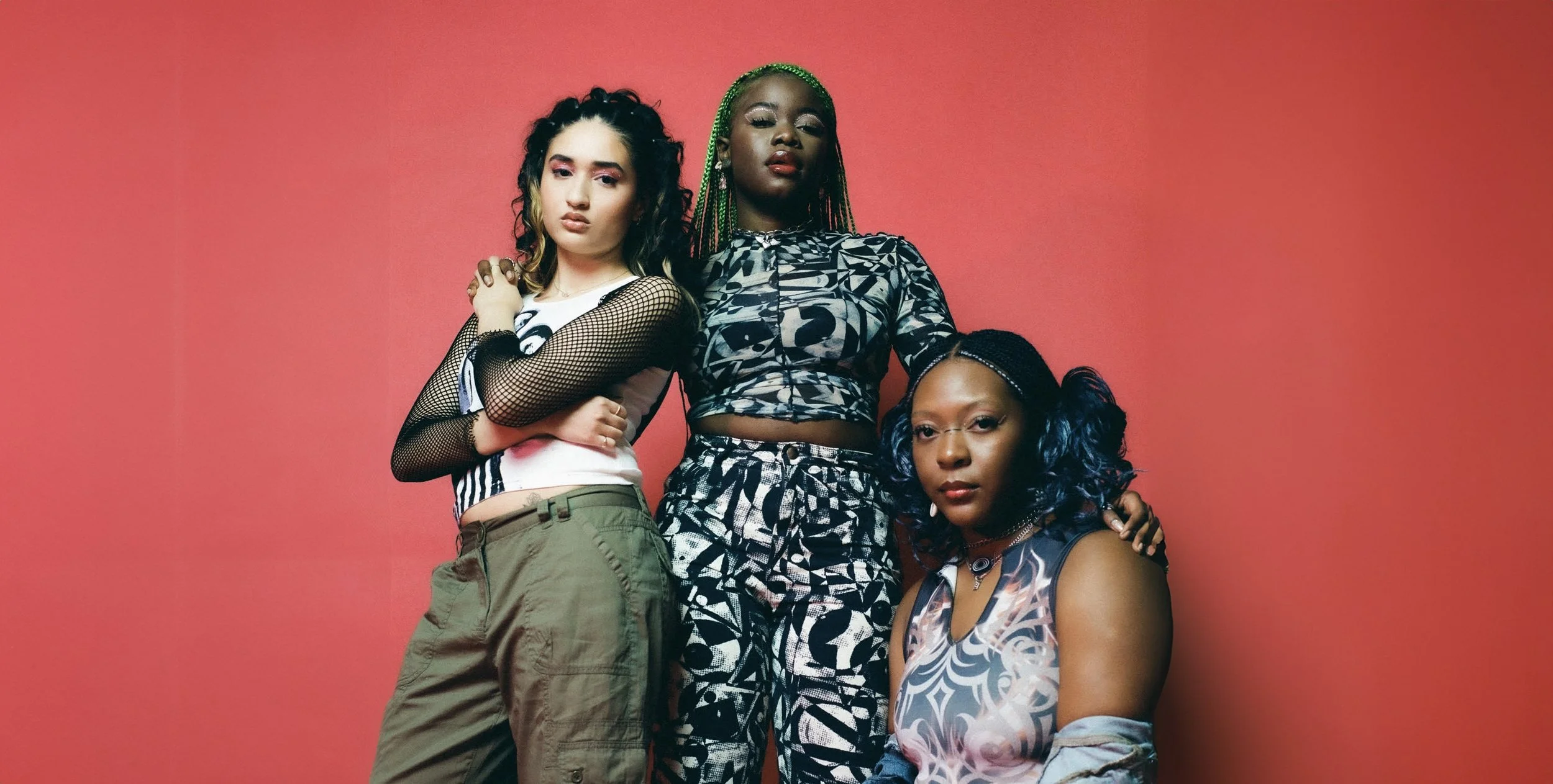 Three young women standing close together against a coral pink background. One is wearing a white and black mesh sleeve top with cargo pants, another in a patterned cropped long-sleeve top and matching pants, and the third in a patterned sleeveless d