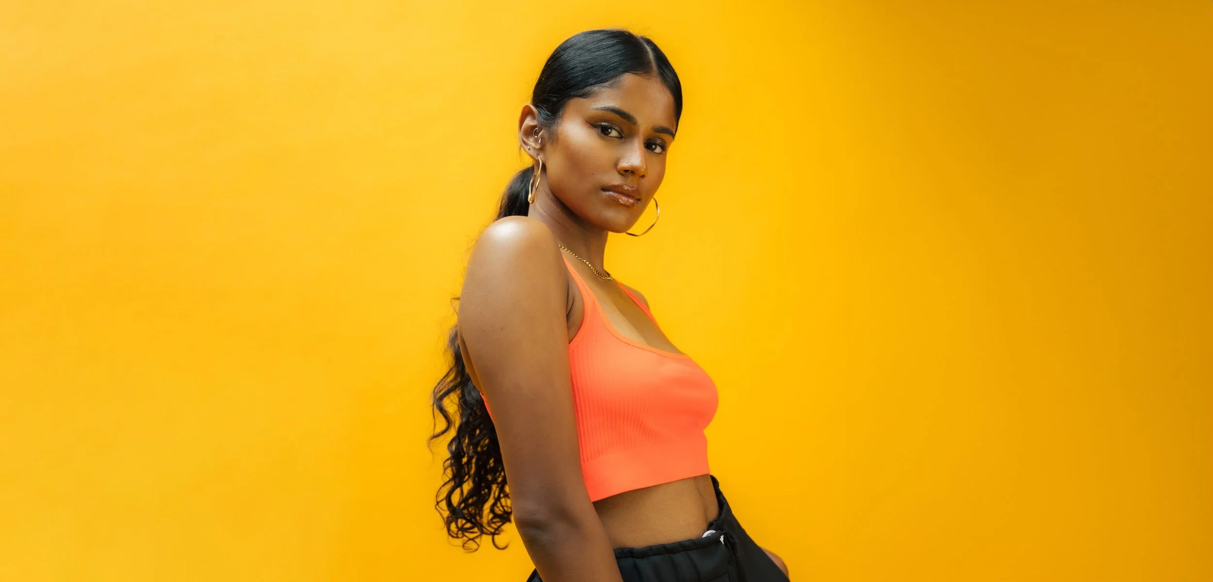 Young woman with long dark hair in a ponytail, wearing hoop earrings, a gold necklace, an orange sleeveless top, and black pants, standing against a yellow background.
