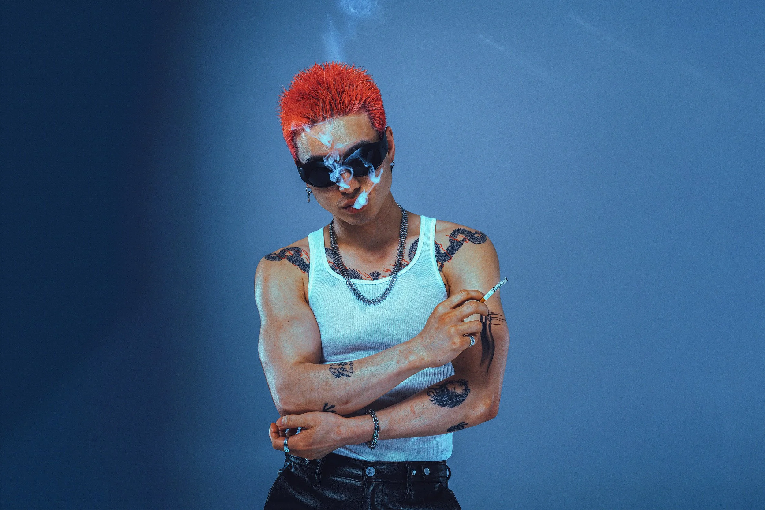 A person with bright red hair wearing dark sunglasses, a white tank top, and tattoos. They are smoking a cigarette with smoke nearby, and are set against a blue background.