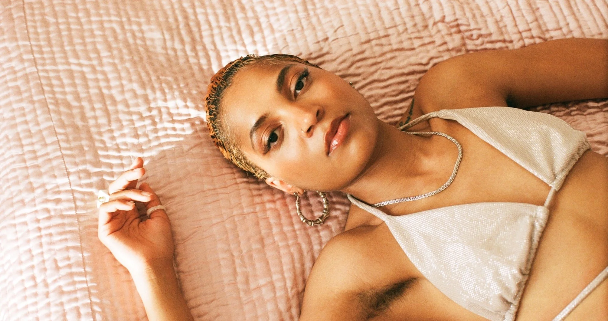 A woman with blonde hair lying on a pink quilted bed, wearing a shiny silver top, hoop earrings, and a silver necklace.