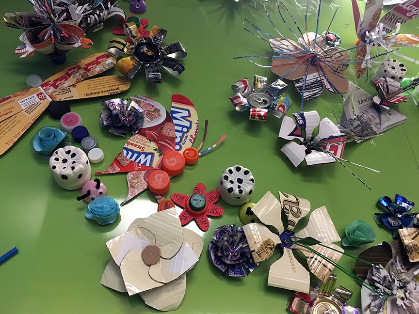 Materials included plastic bottles and lids, aluminum cans, cardboard, newspaper, junk mail catalogs, office waste paper, and printed food boxes. The variety of designs and methods for making the art objects was unlimited. Each item is individually attached to the wall with magnets.