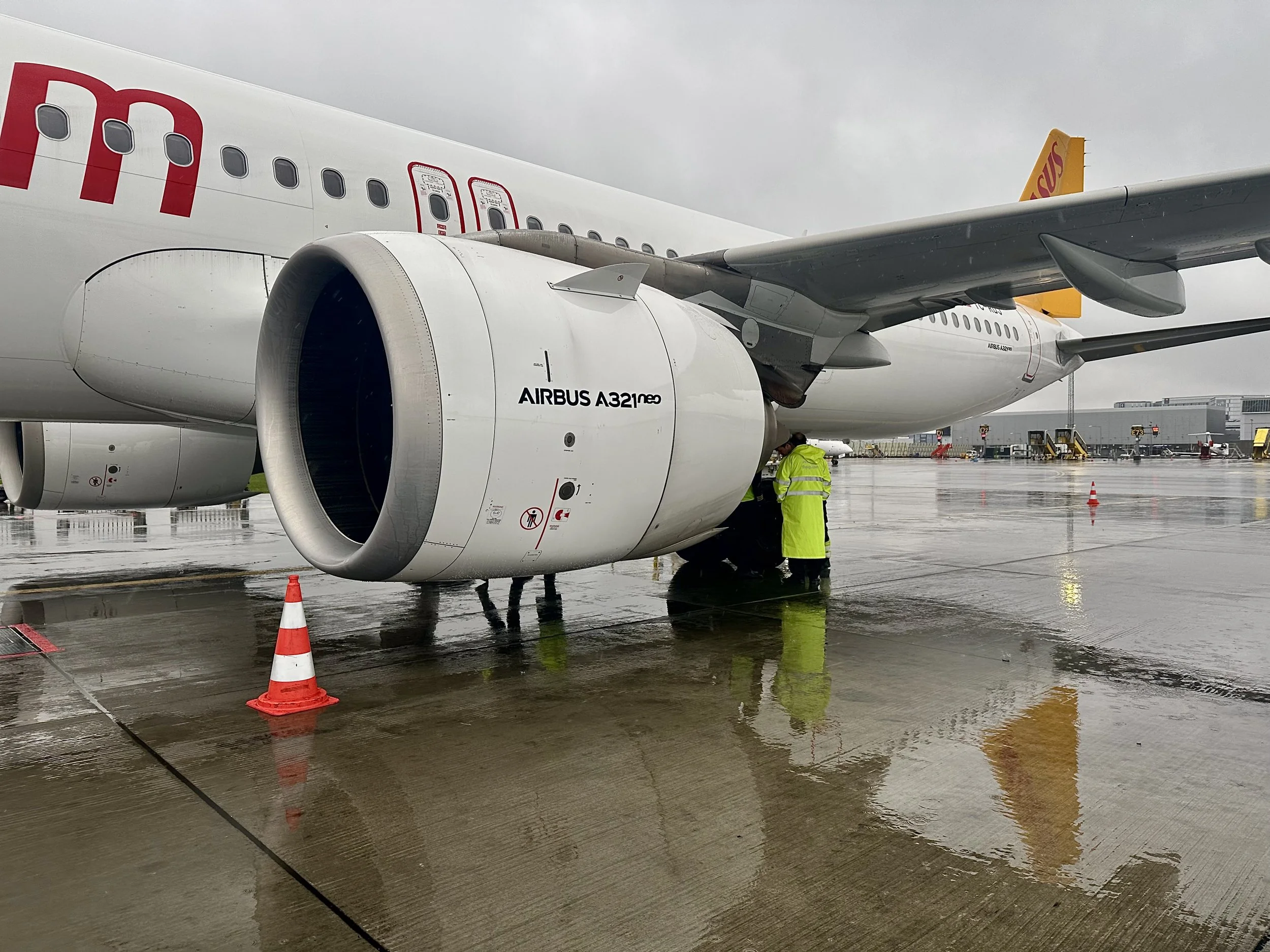 A close-up view of an Airbus A321 aircraft on wet tarmac, with ground crew members in reflective yellow safety vests working near the plane, and orange traffic cones placed around.