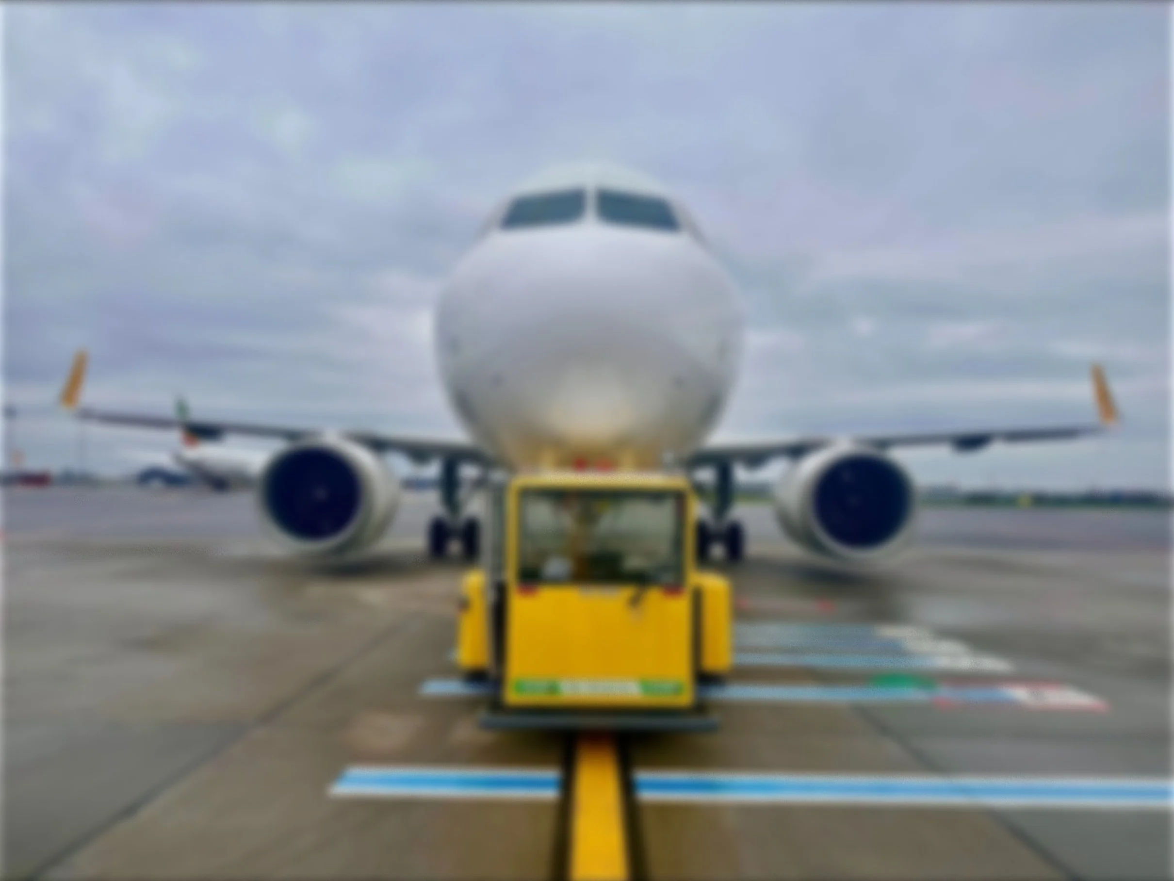 Front view of a white commercial airplane on the tarmac, with a yellow tug in front of it. Overcast sky.