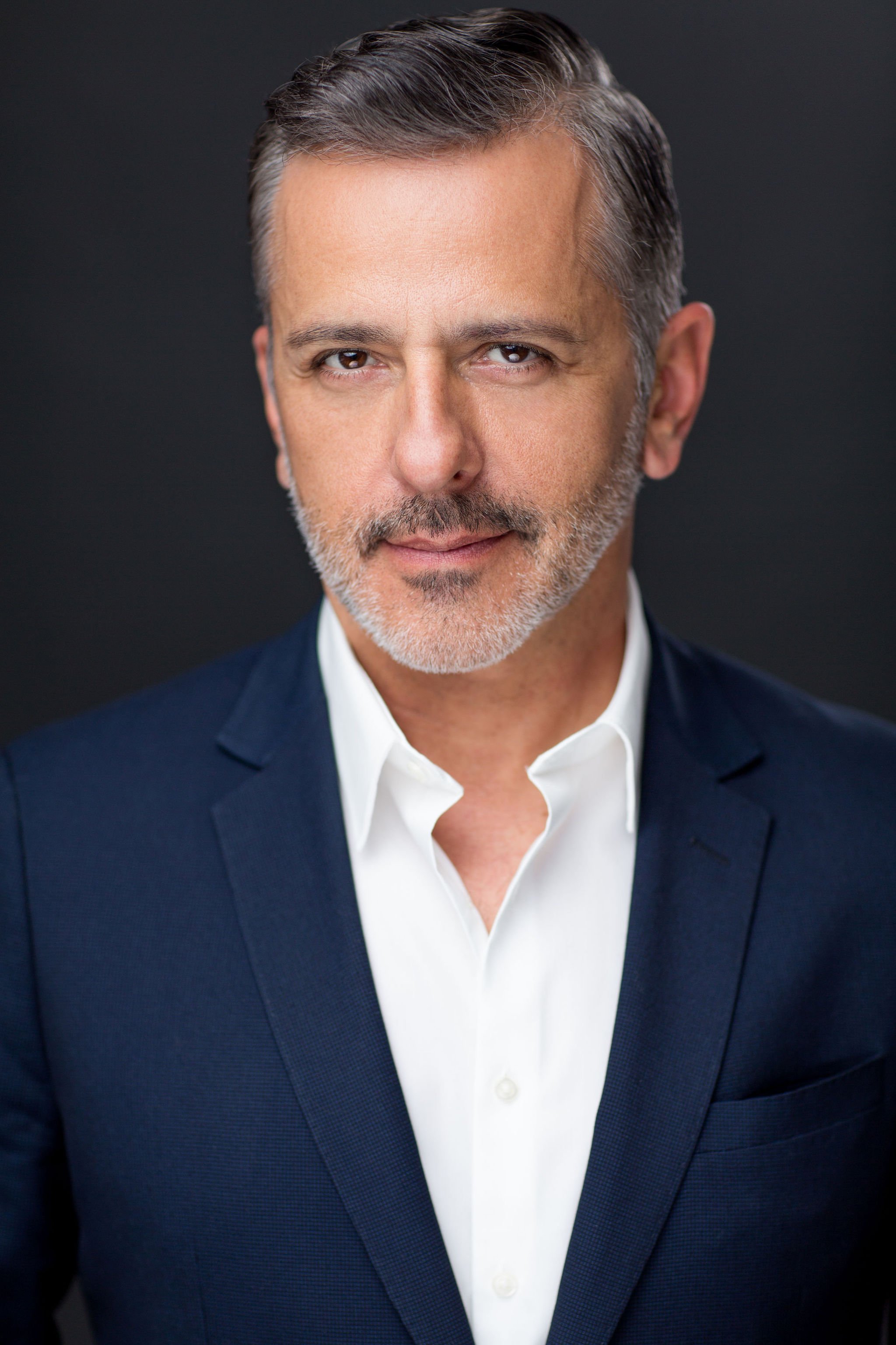 Professional headshot of a man with a salt-and-pepper beard wearing a navy suit against a dark background.