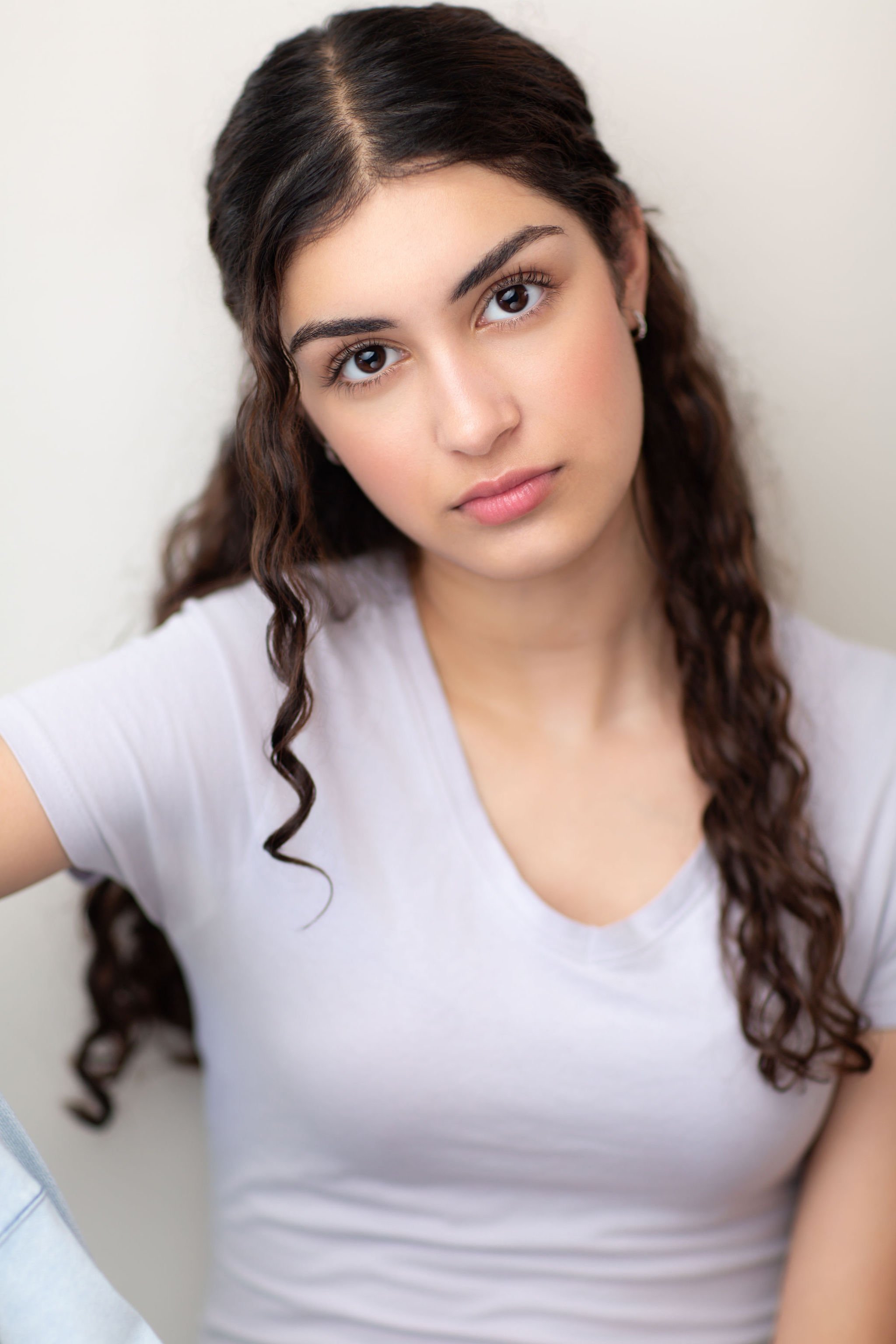 Actor headshot of a young woman with long curled hair wearing a white top against a pale background.