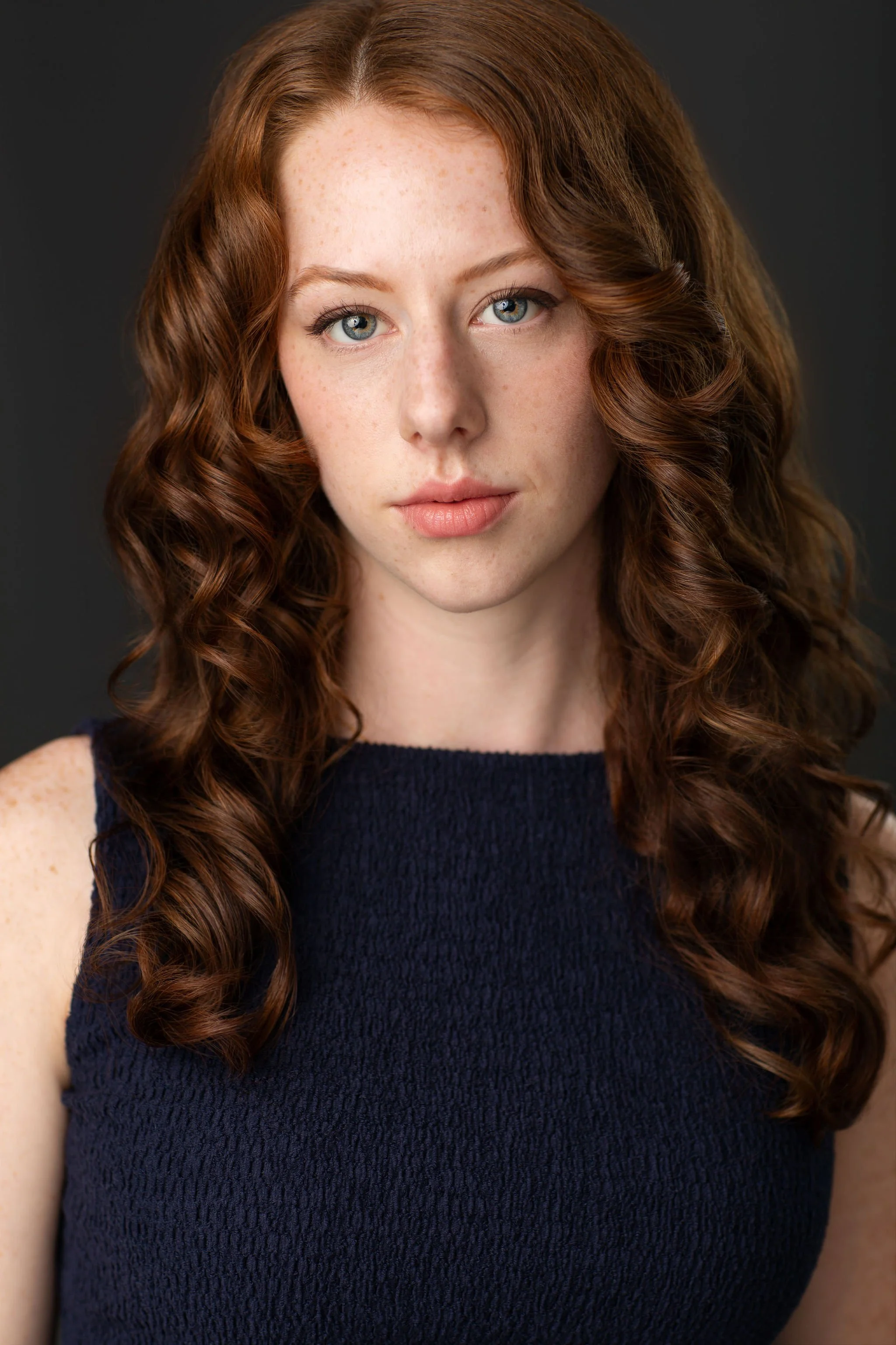 Actor headshot of a red-haired woman wearing a navy sleeveless top against a dark background.
