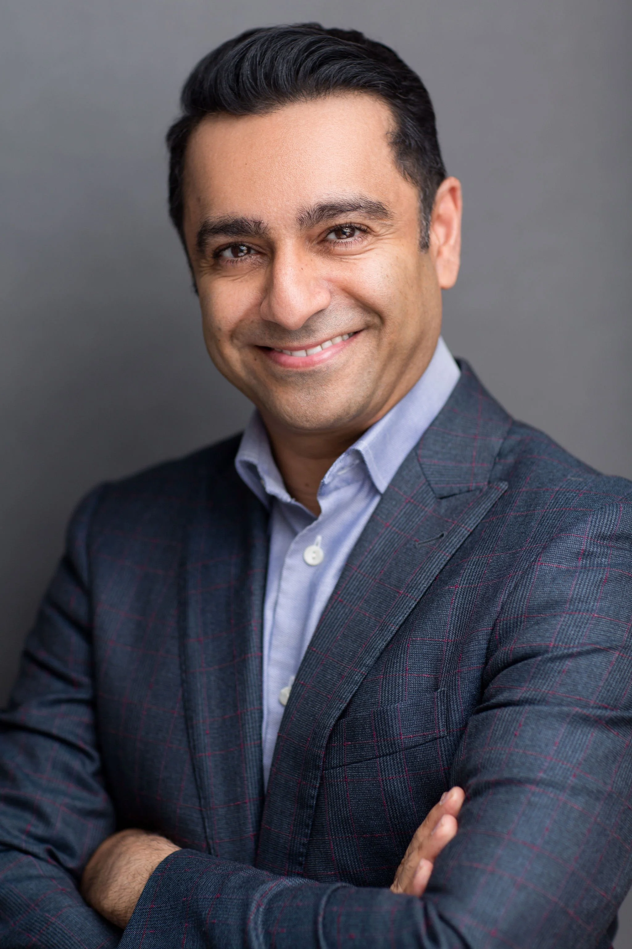 Professional headshot of a smiling man in a grey suit with his arms crossed against a grey background.