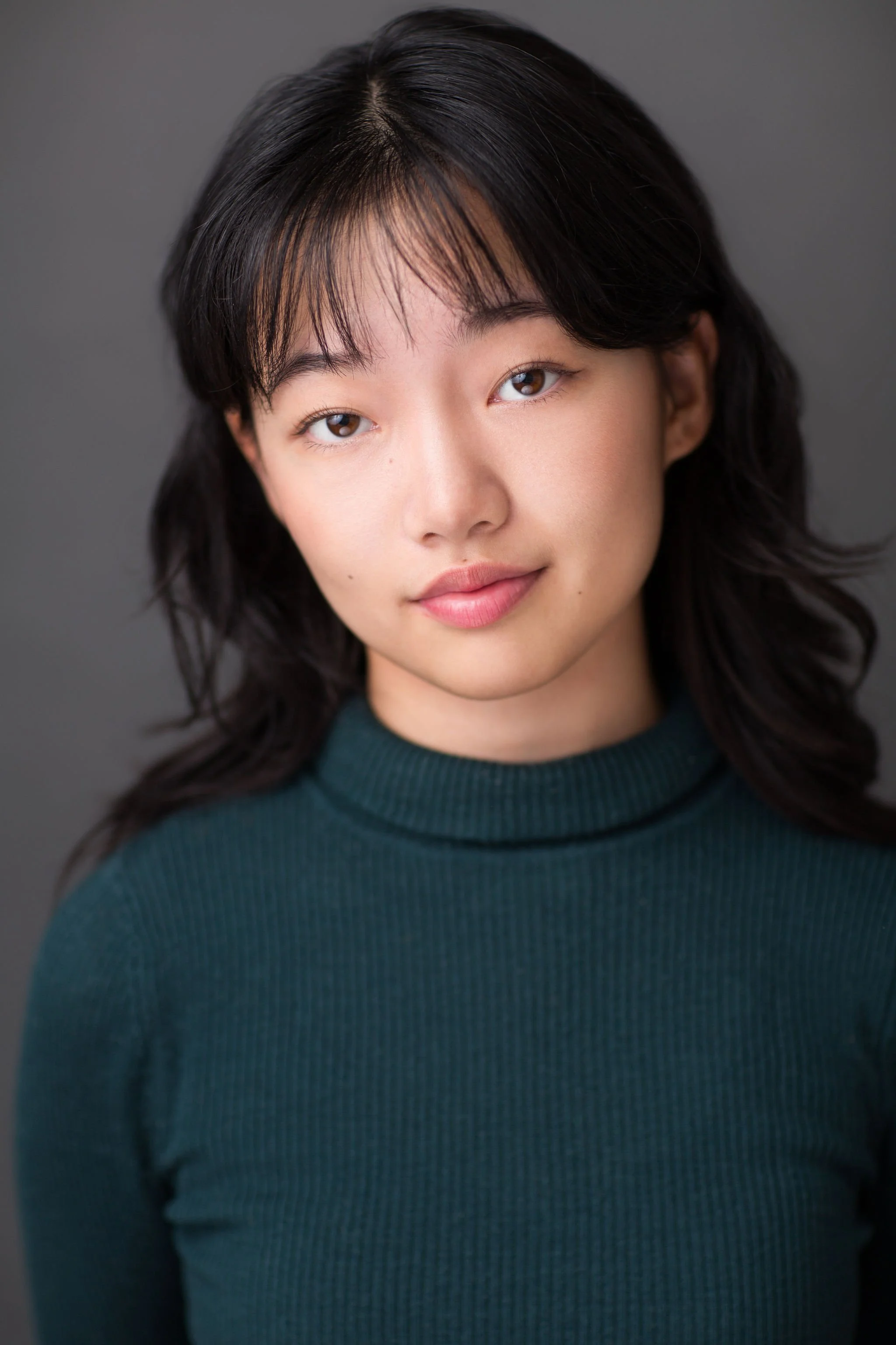Actor headshot of a young woman with dark wavy hair wearing a green turtleneck against a grey background.