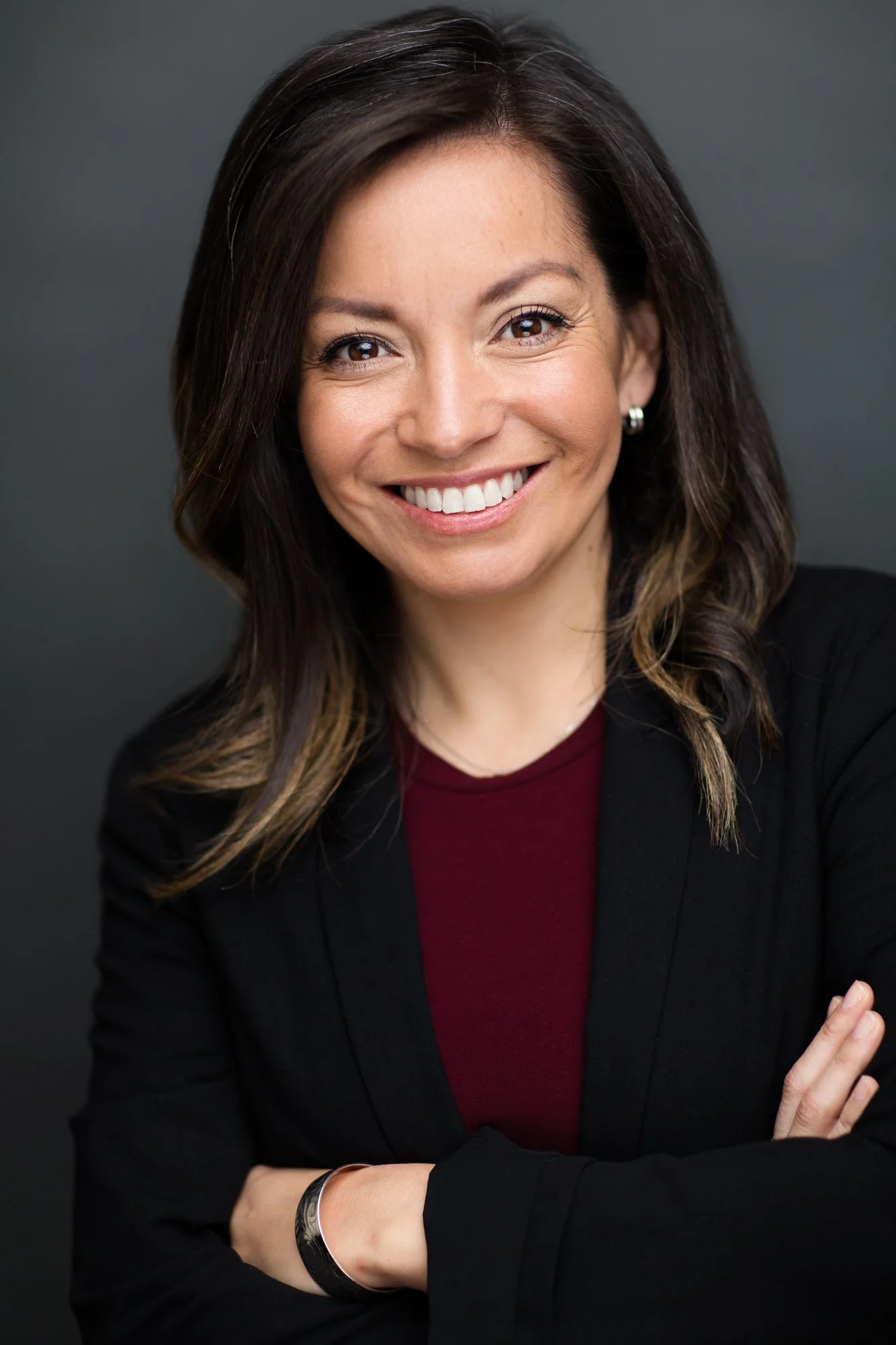 Business professional commercial headshot of a smiling woman in a black blazer with her arms crossed against a dark background.