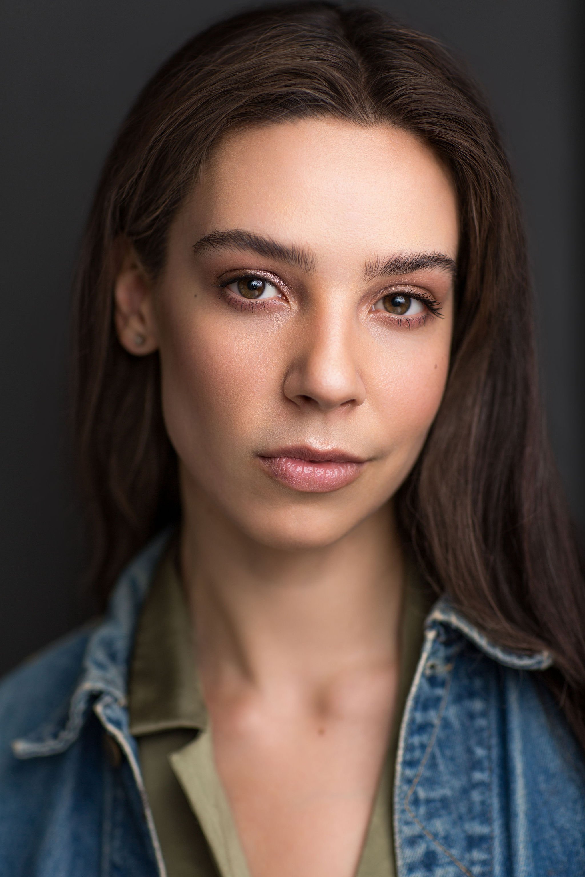 Actor headshot of a brunette woman wearing a denim jacket against a dark studio background.