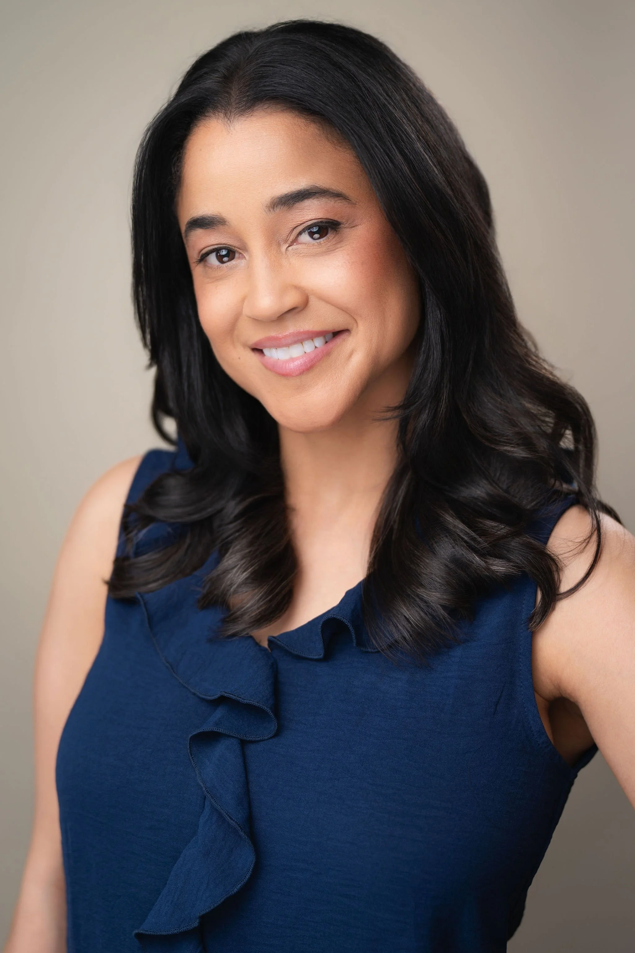 Professional commercial headshot of a smiling woman in a navy sleeveless blouse against a warm neutral background.