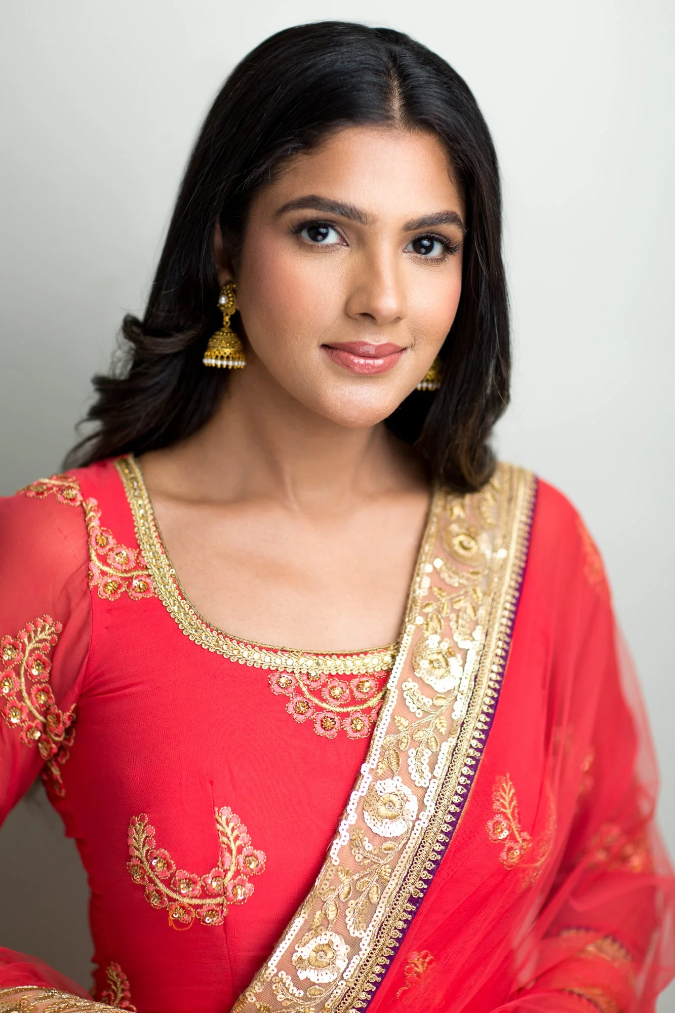 Portrait of a woman wearing a red traditional South Asian outfit with gold trim against a pale background.