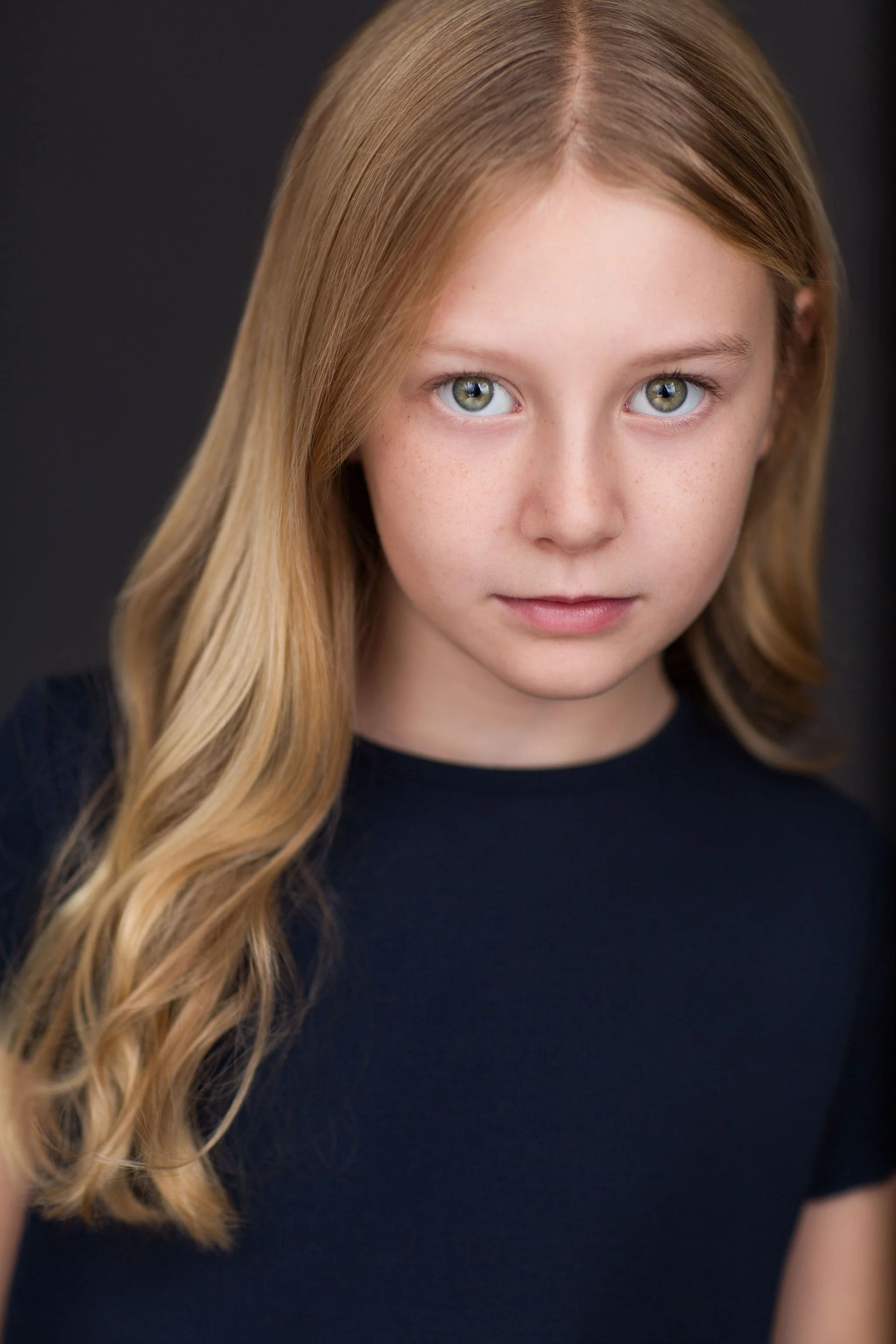 Child actor headshot of a blonde girl wearing a navy top against a dark studio background.