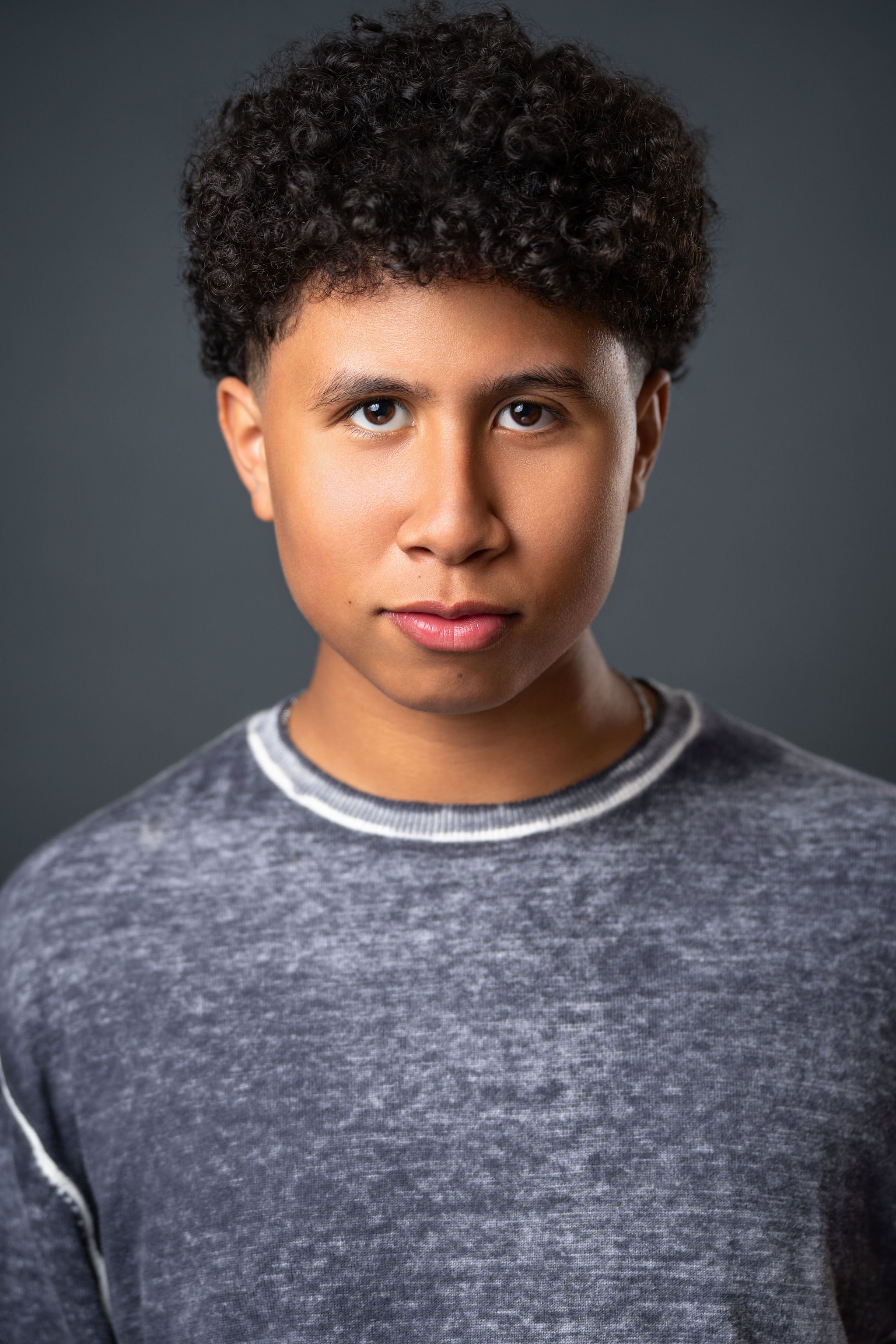 Actor headshot of Casey Trotter in a grey sweater against a dark grey studio background.