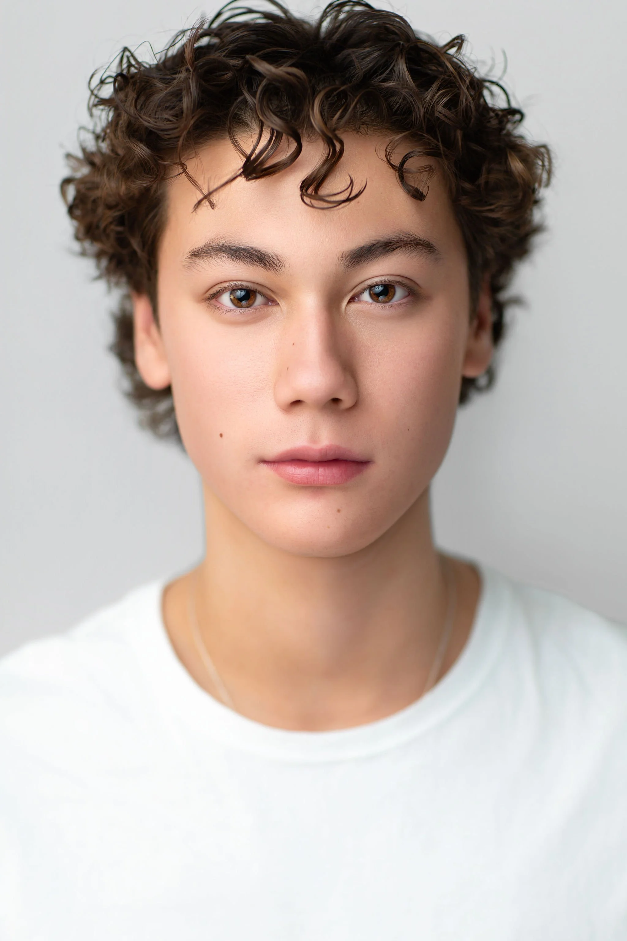 Teen actor headshot of a boy with curly hair wearing a white T-shirt against a light grey background.