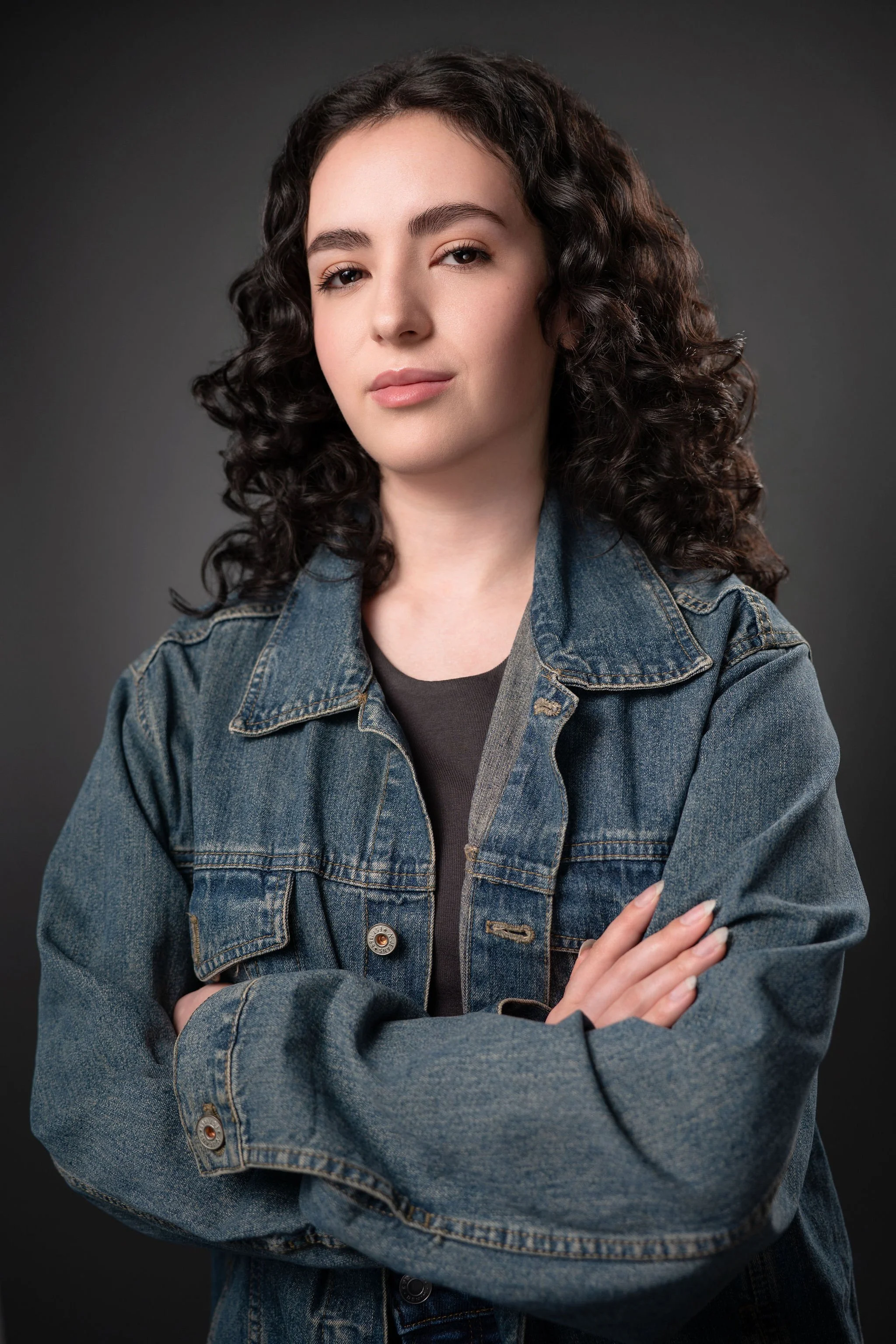 Actor portrait of a curly-haired woman in a denim jacket with her arms crossed against a dark background.