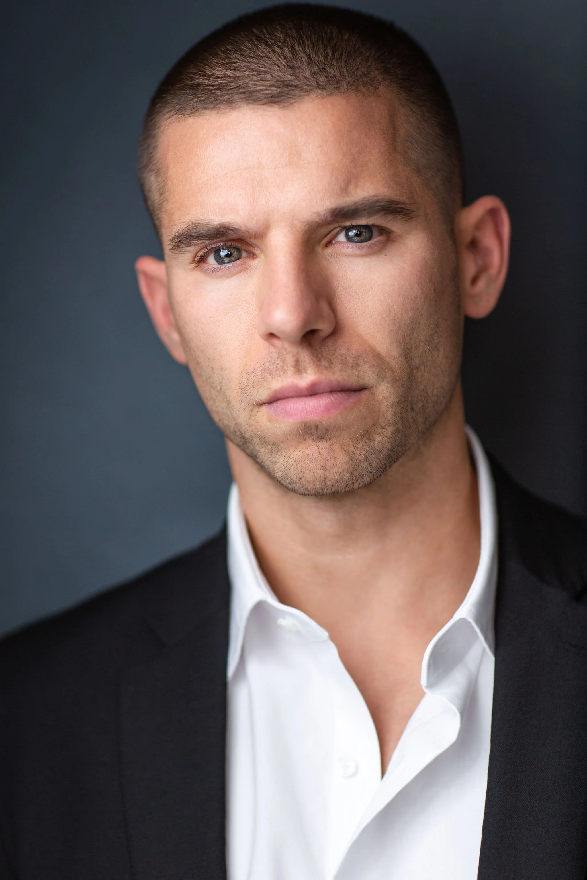 Actor headshot of a man with a buzz cut wearing a black blazer and white shirt against a dark background.