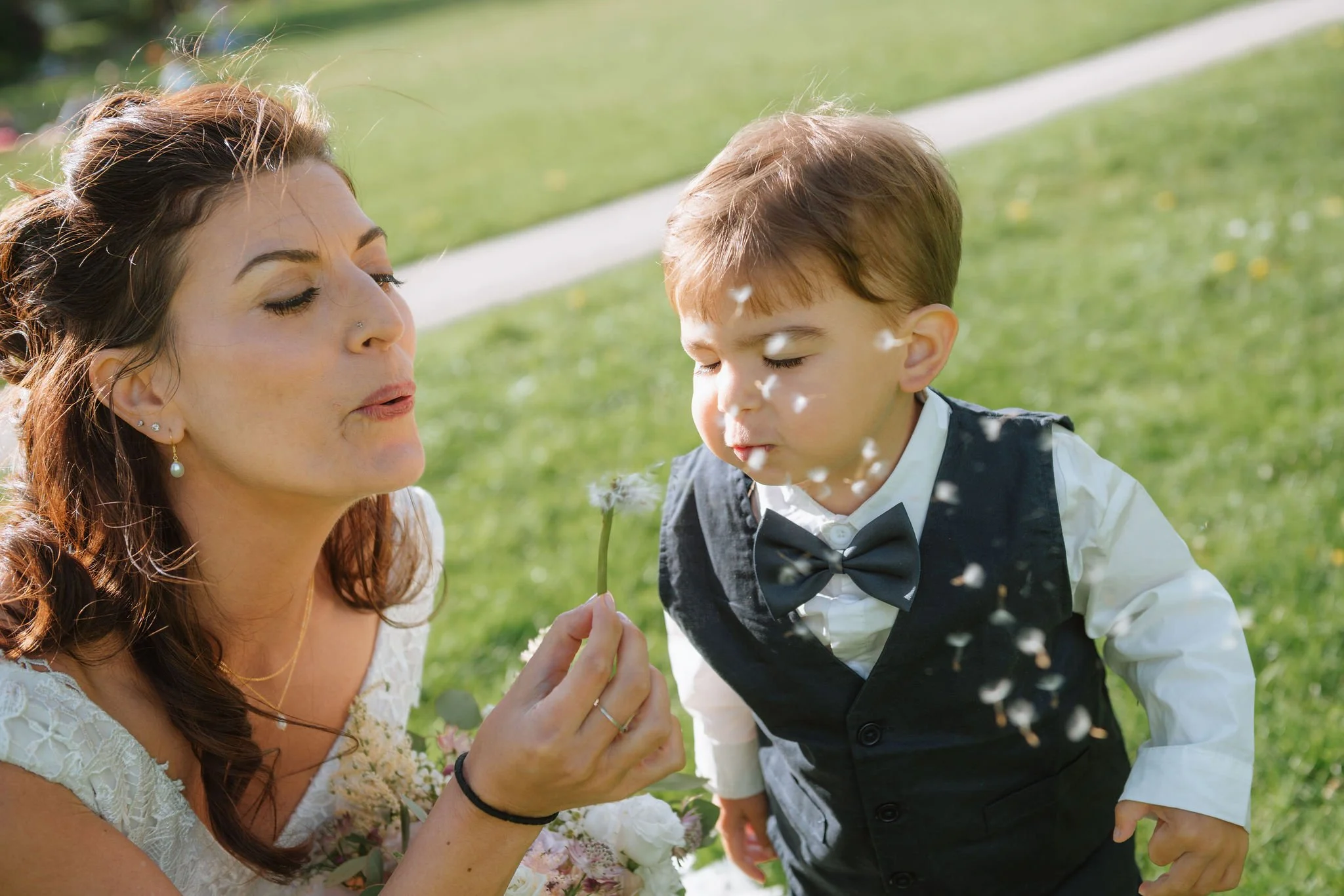 Bride blowing a dandelion with a young ring bearer during an outdoor wedding celebration.