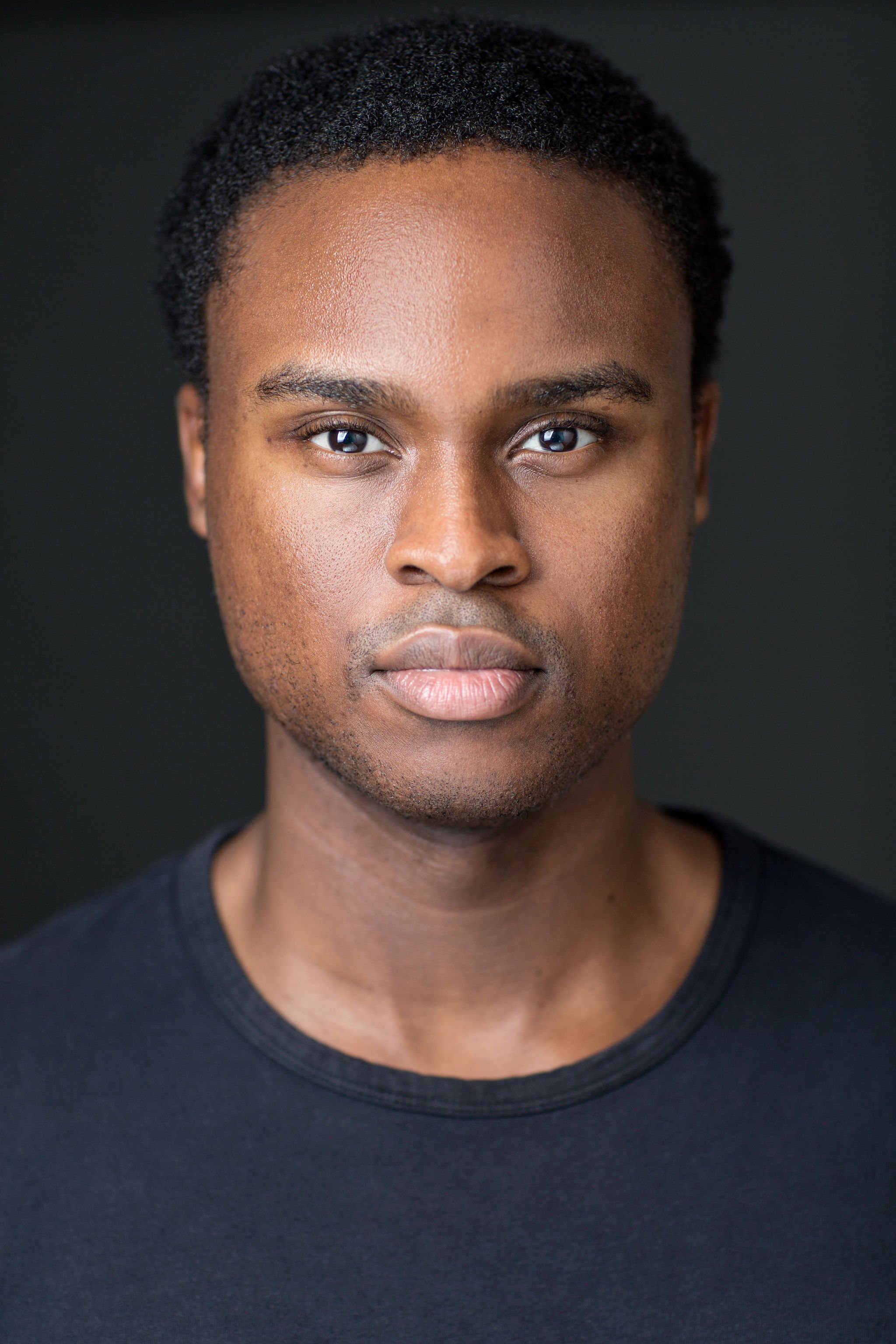 Actor headshot of a man with close-cropped hair wearing a black T-shirt against a dark grey background.